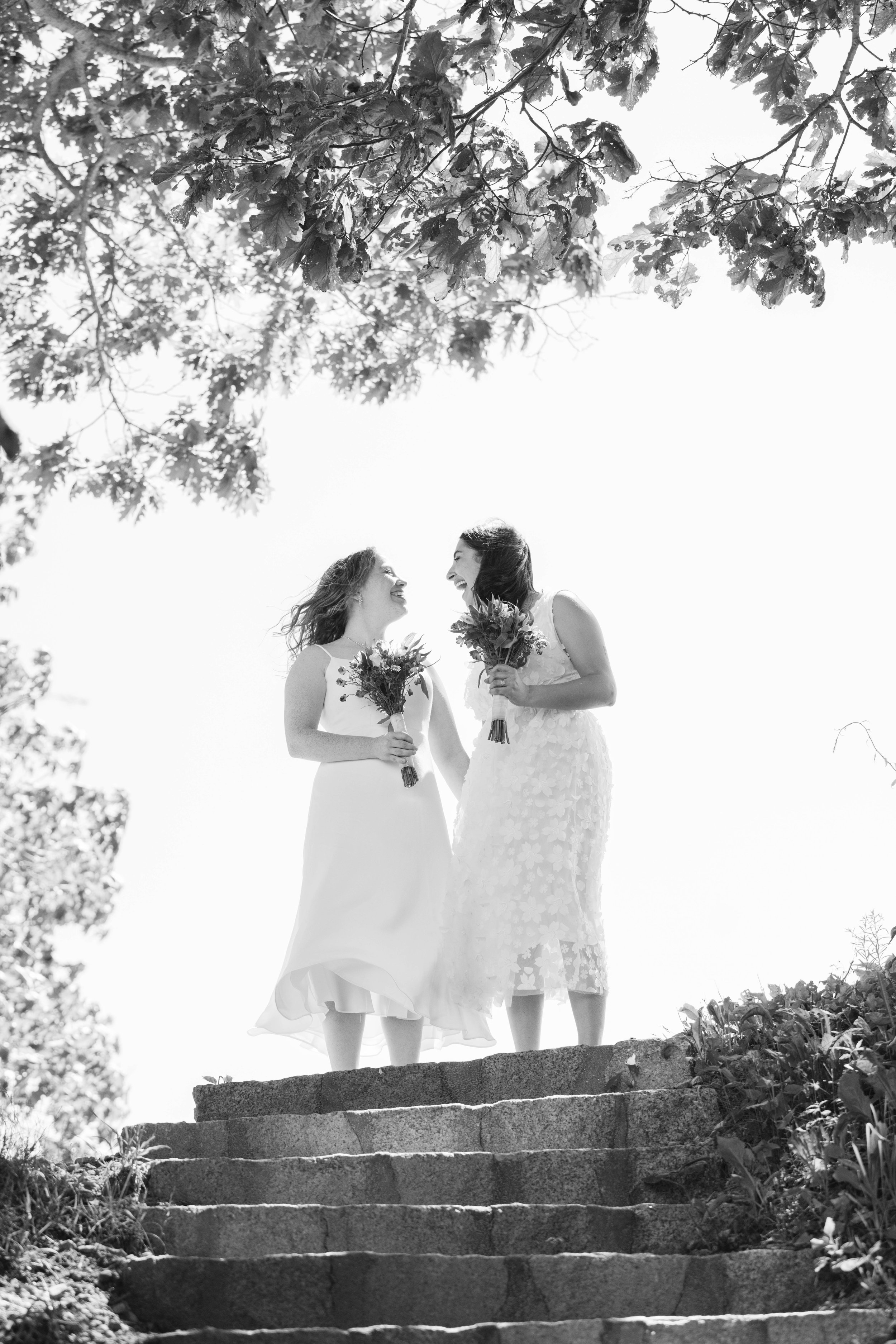 Two women in wedding dresses holding bouquets, standing on stone steps outdoors, smiling and looking at each other, surrounded by trees.