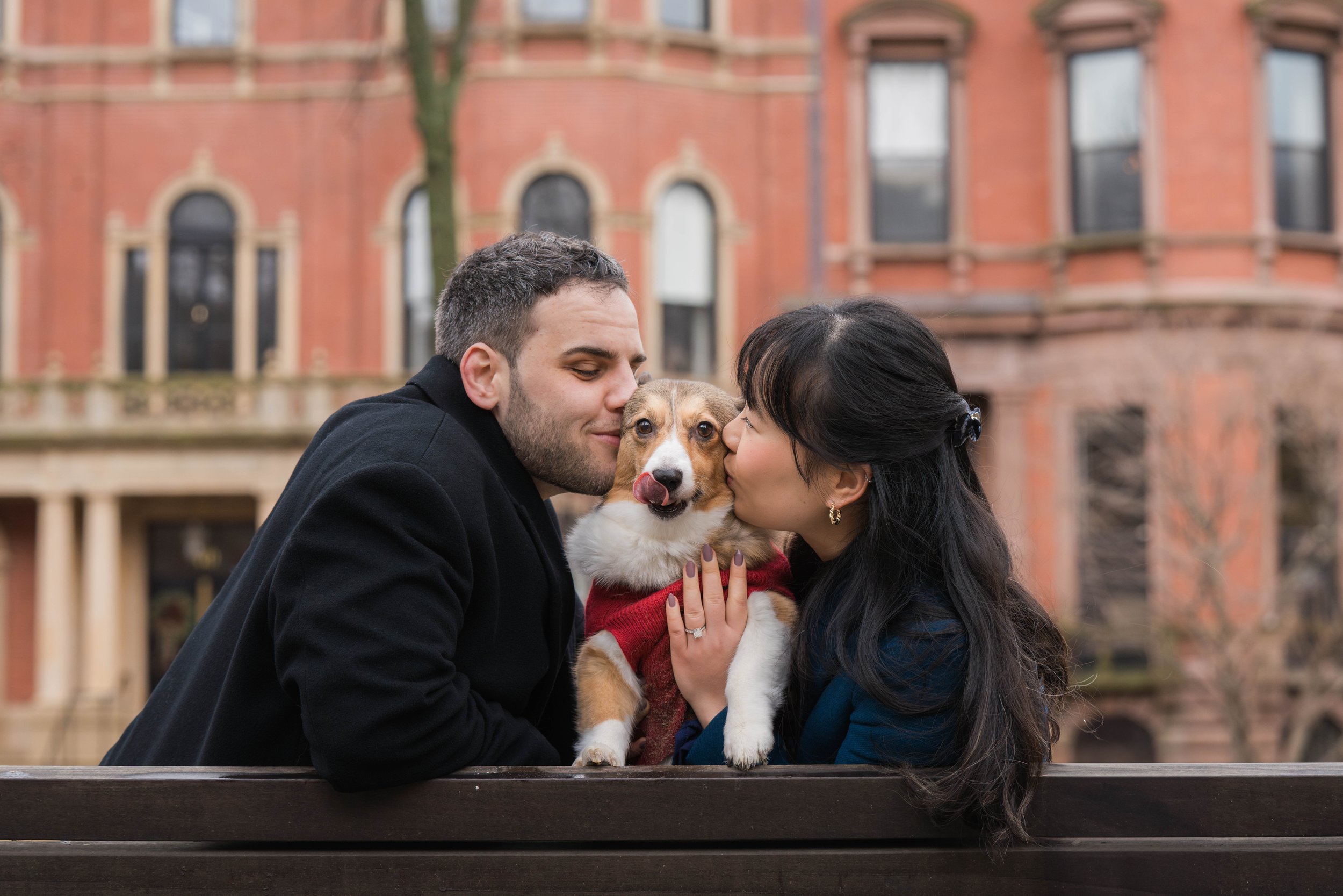 A couple with their dog outdoors in front of a red brick building, sharing a kiss while holding the dog