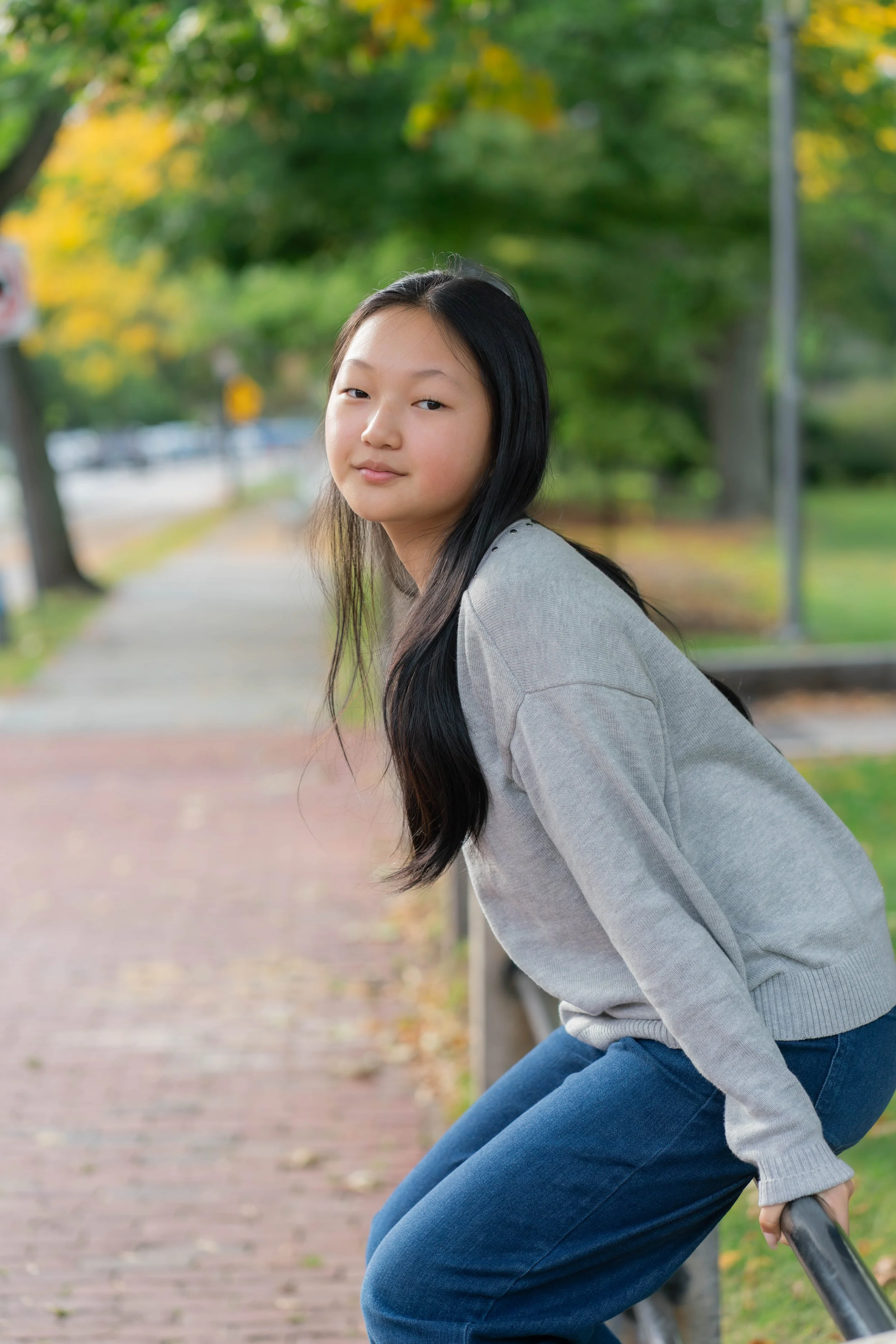 A young woman with long black hair, wearing a grey sweatshirt and blue jeans, leaning on a metal railing outdoors during daytime with trees and a pavement in the background.