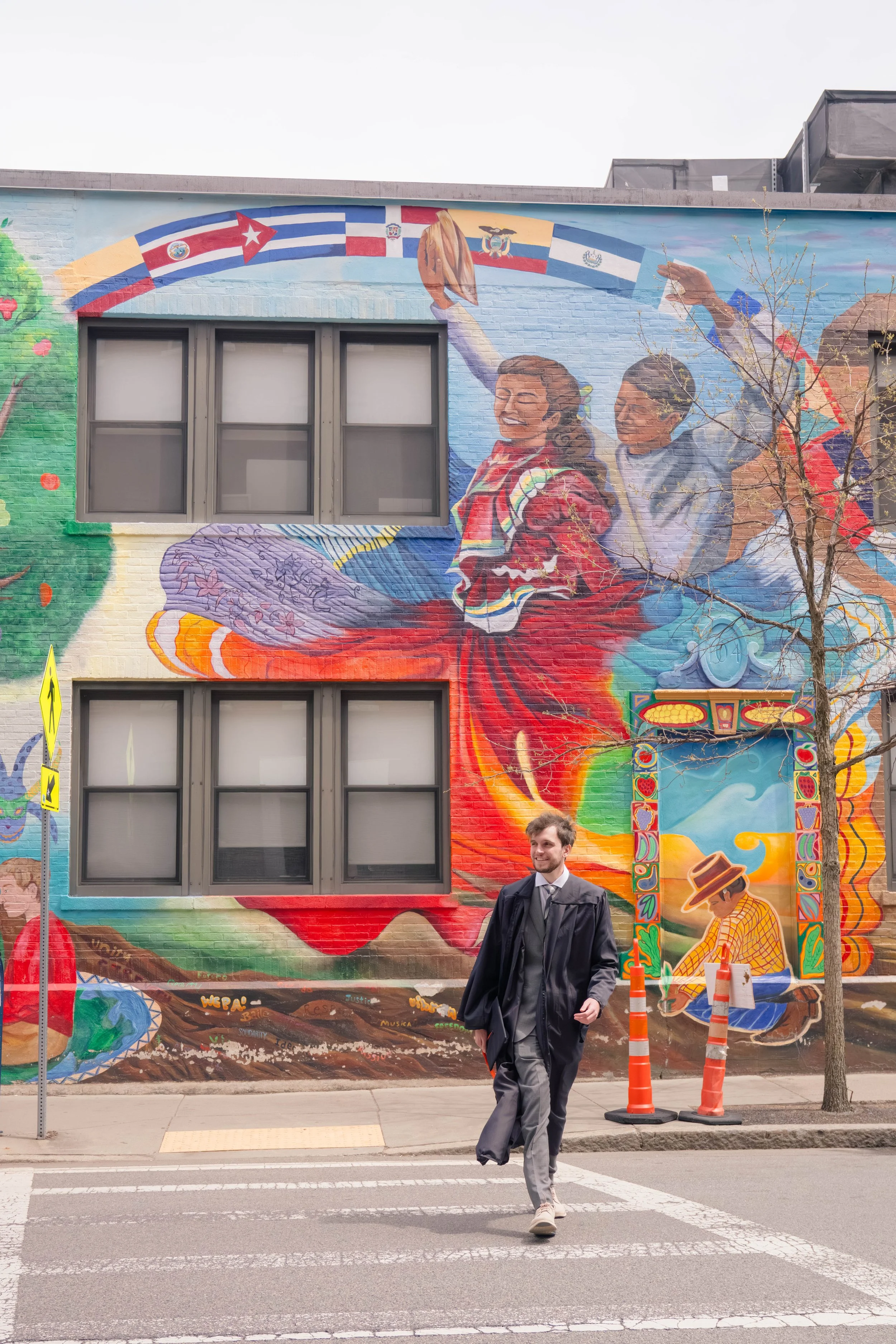 A man in gray pants, a white shirt, a black tie, and a black graduation gown walking across a crosswalk in front of a colorful mural on a building wall. The mural depicts smiling women, a tree, and various flags including Cuba, Ecuador, and Paraguay,