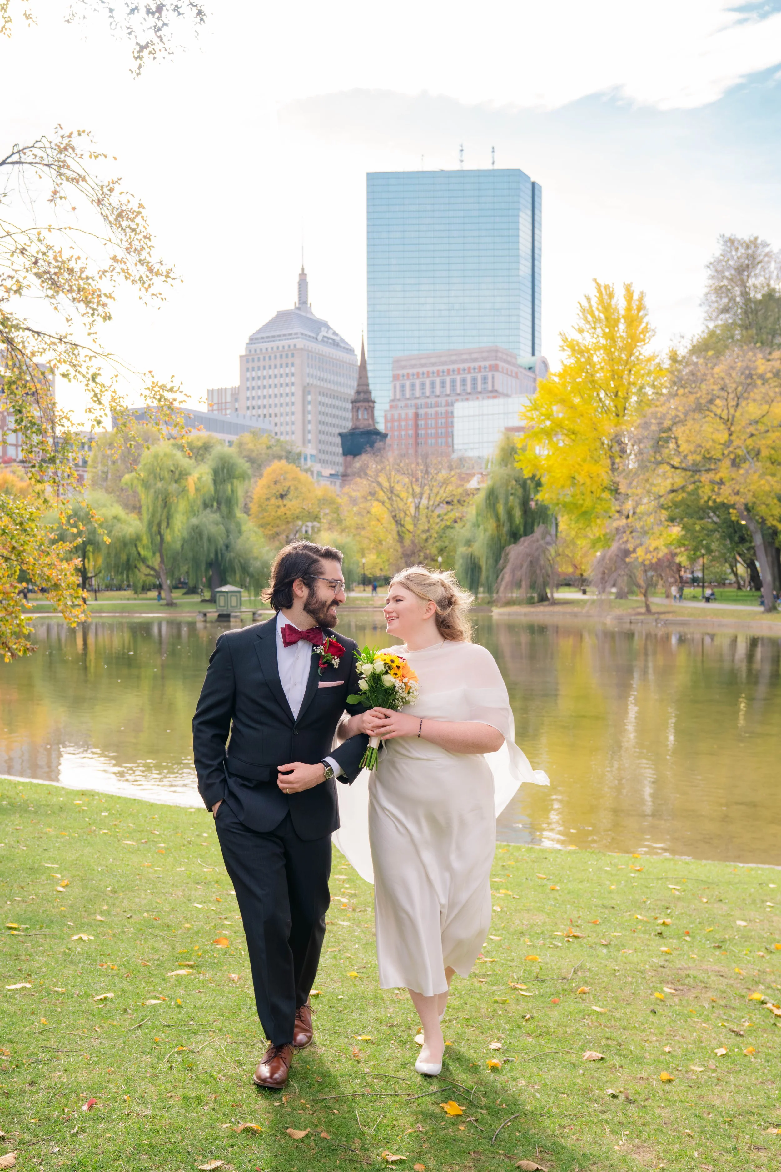 A bride and groom walking arm in arm by a pond in a park with autumn trees and city skyscrapers in the background.