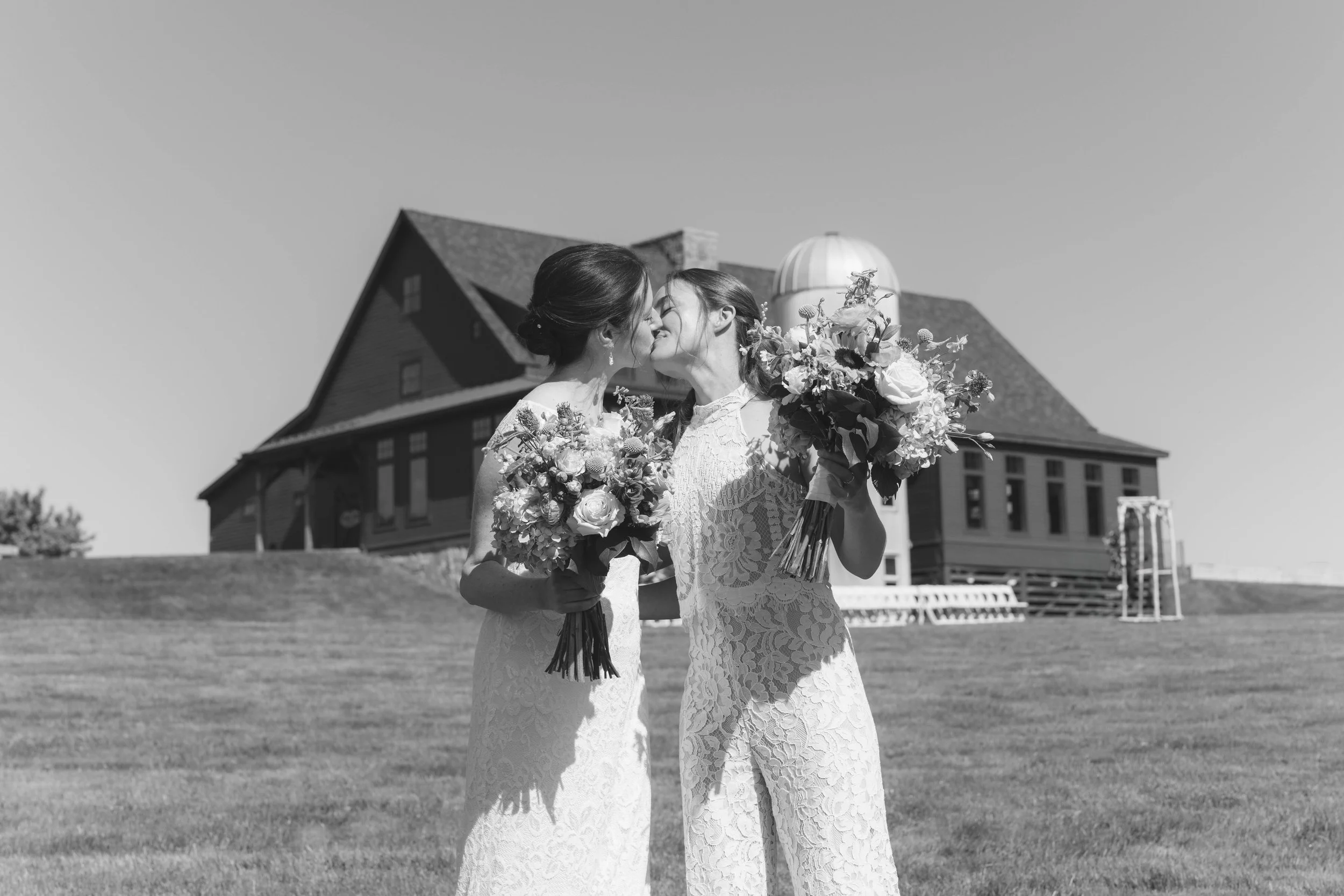 Two brides in lace wedding dresses sharing a kiss outdoors, each holding a large bouquet of flowers, with a large house in the background.