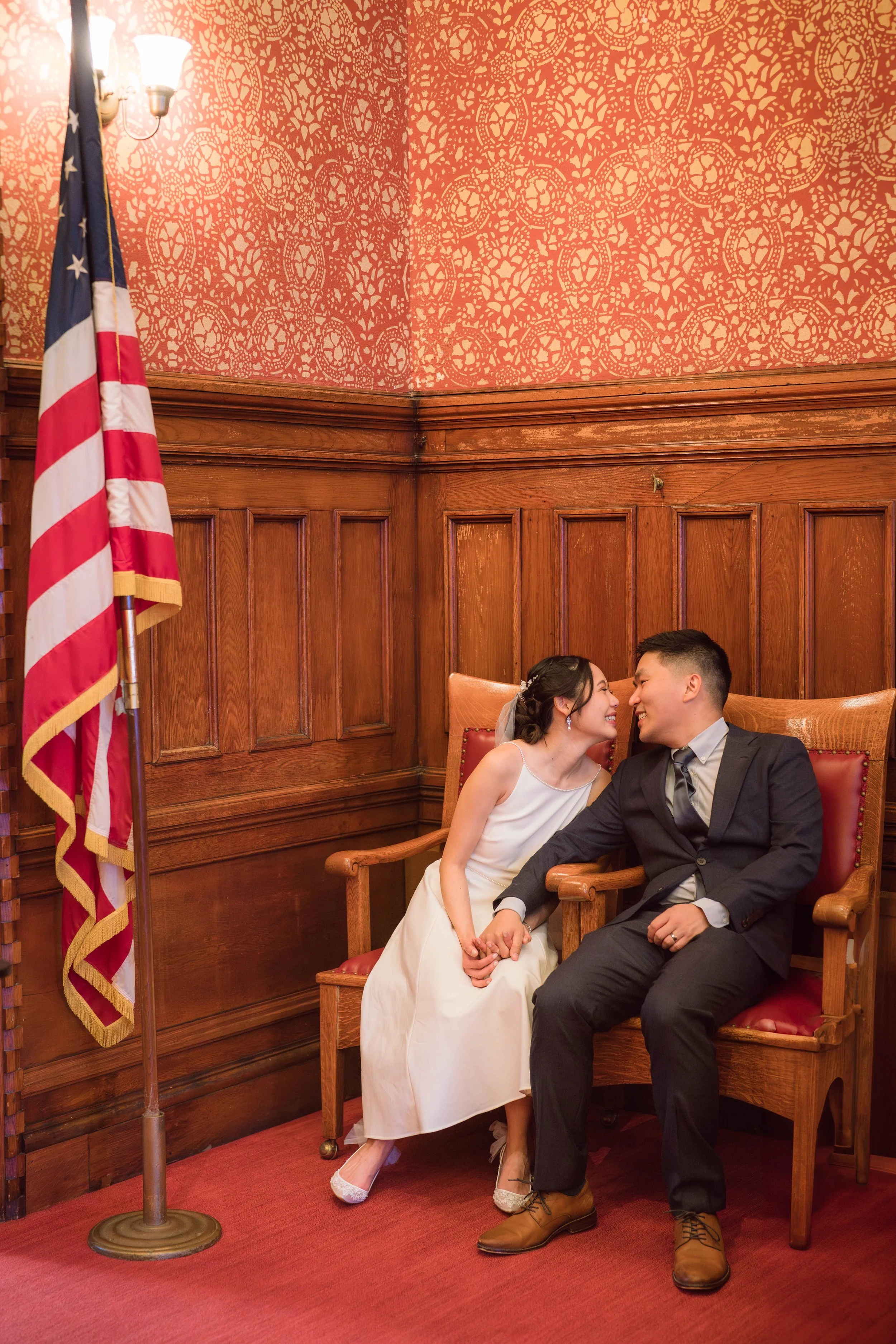 A couple dressed in wedding attire sitting on a bench, holding hands, in a wood-paneled room with red carpet, patterned wallpaper, and an American flag.