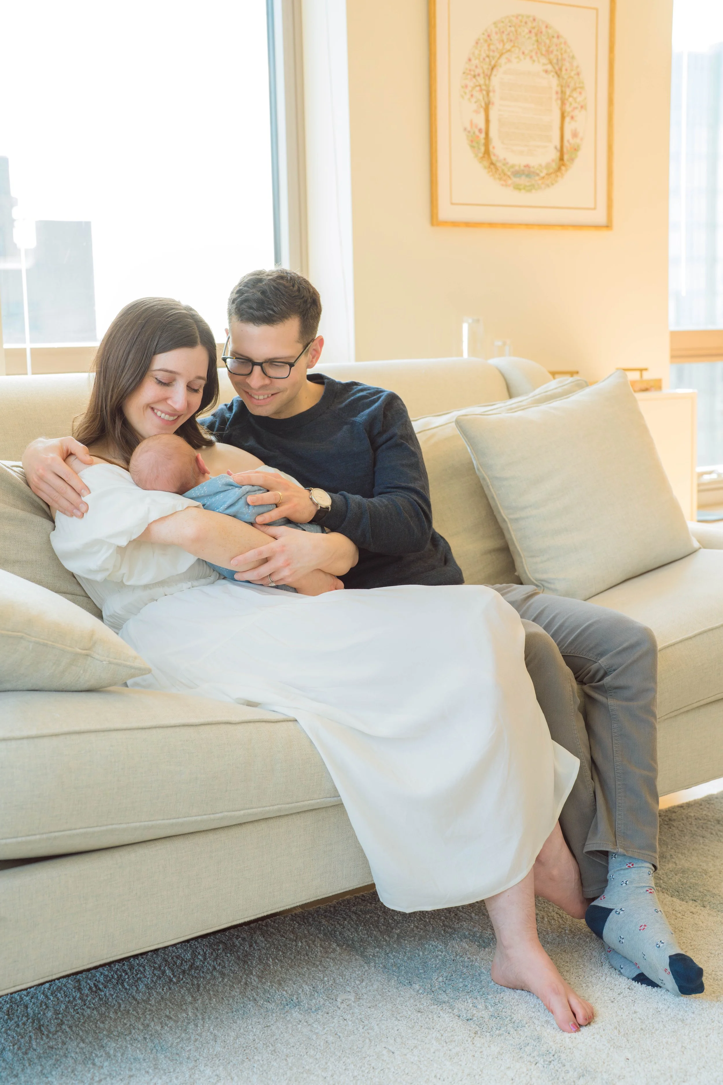A happy family sitting on a beige couch in a living room, with a woman holding a newborn baby wrapped in a blue blanket, and a man smiling beside them. The woman is wearing a white dress, the man is dressed in a dark shirt, and a child with socks sit