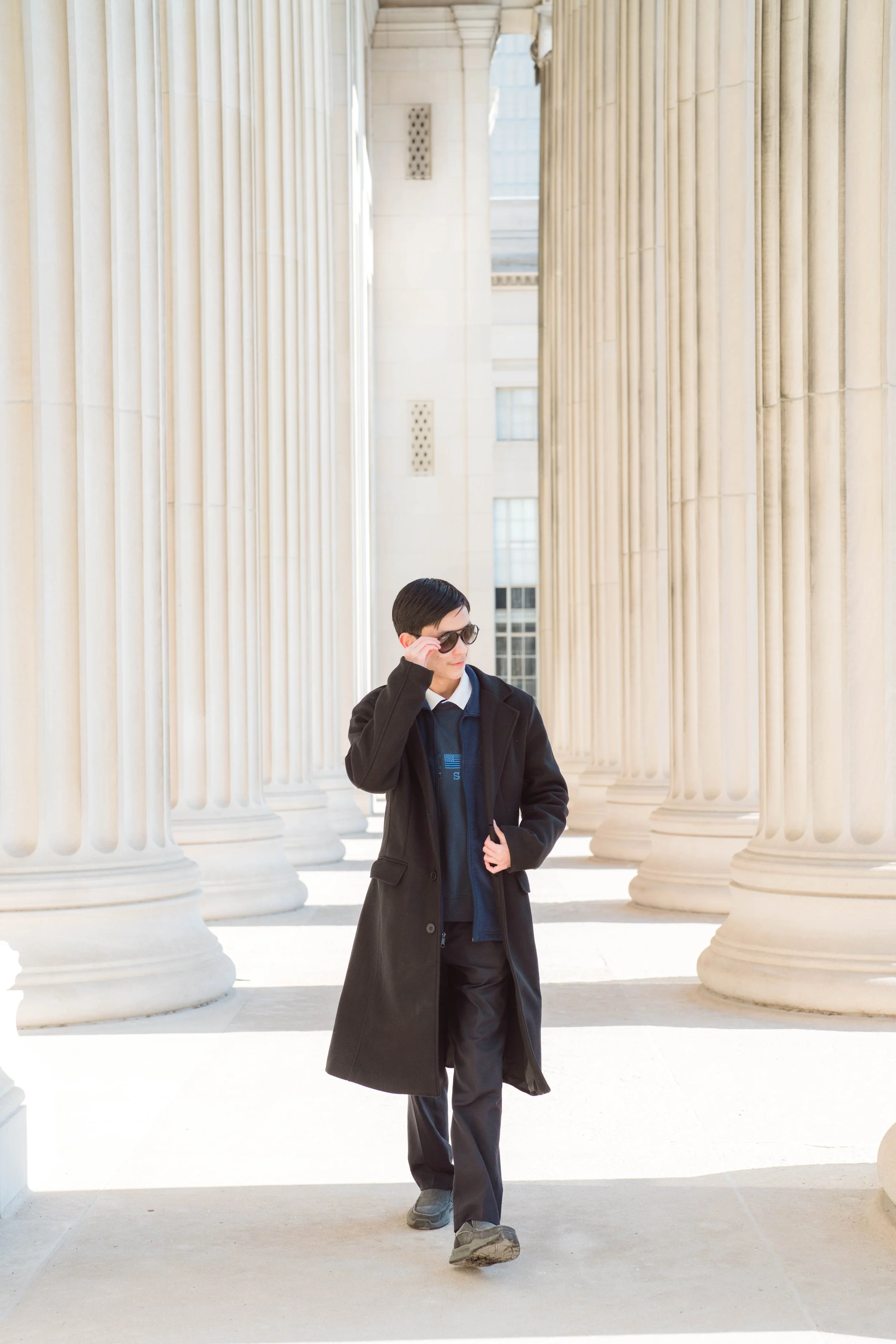 A person with short dark hair, wearing sunglasses, a black coat, and dark pants, walking among large white classical columns outside a government or historical building.