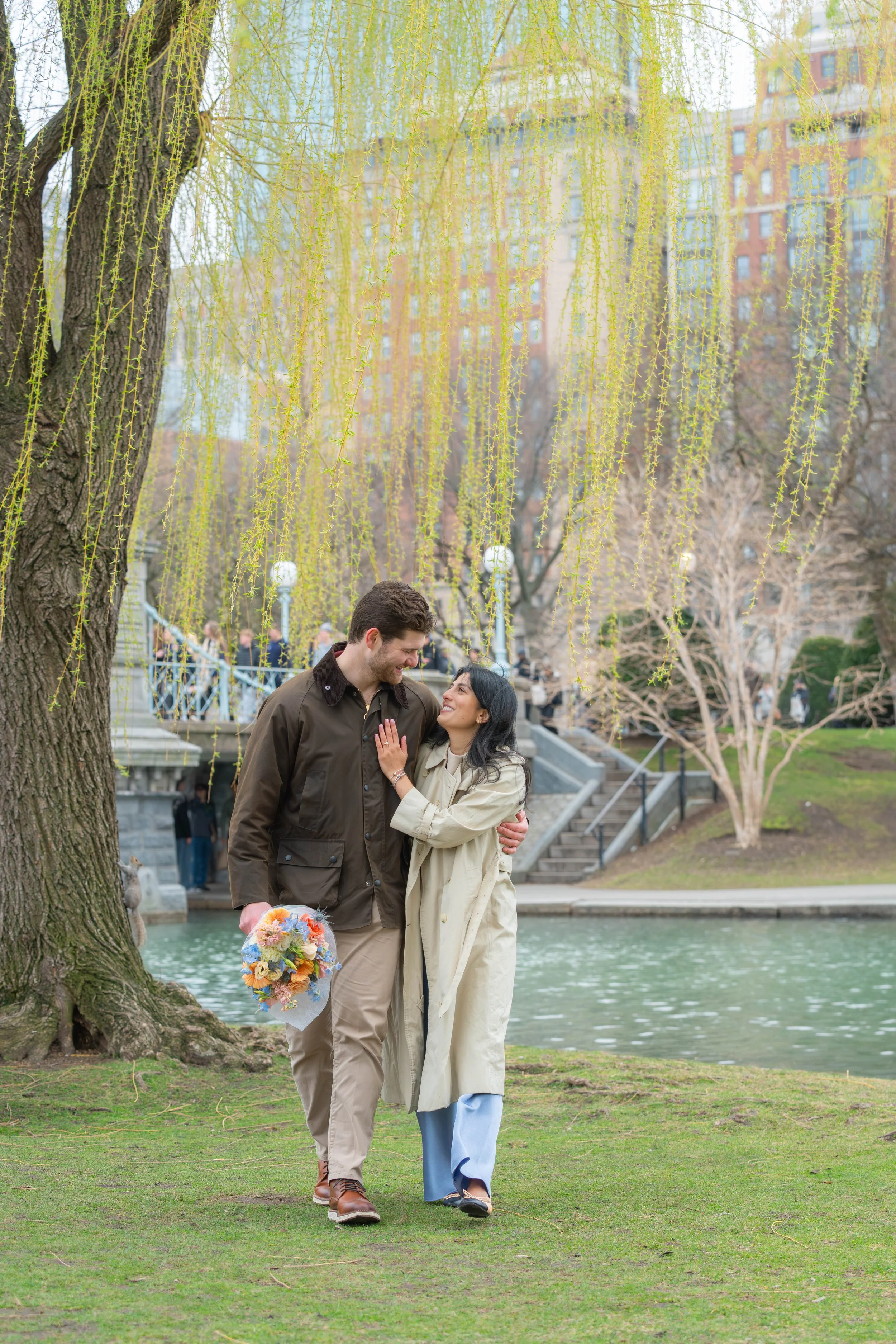 A couple embracing and smiling near a park pond with a large tree with hanging branches and city buildings in the background.