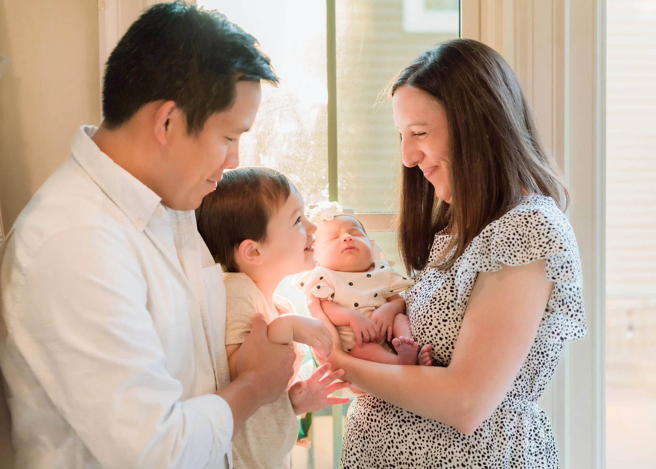 A family of four, including two adults and two children, is gathered near a window. The mother holds a newborn baby, and the father and older child are smiling and looking at the baby.