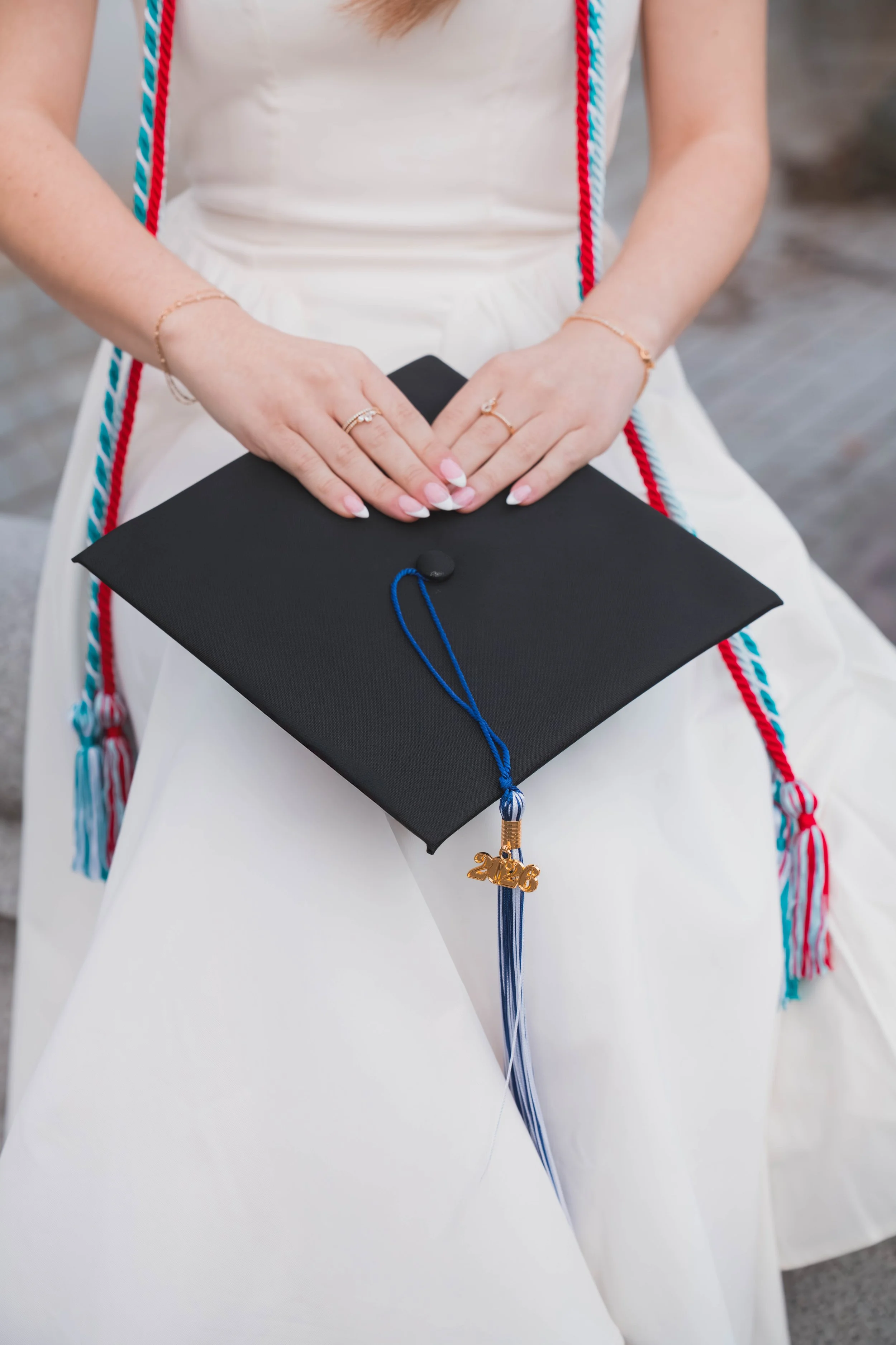 A person wearing a white dress holding a black graduation cap with a blue tassel and a gold '2023' charm, with cords around their neck and rings and bracelets on their hands.