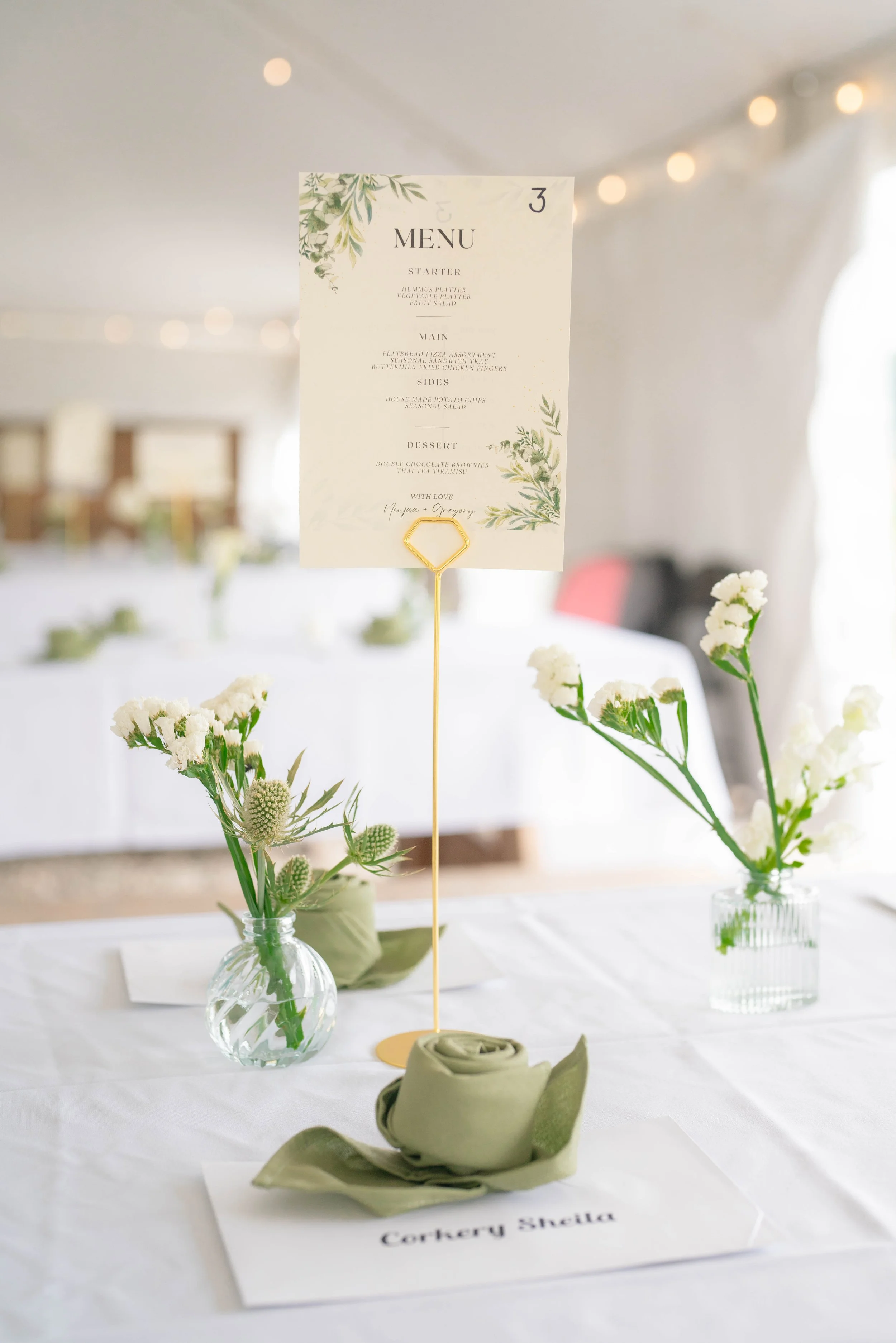 Table centerpiece with white flowers in a small glass vase, a place card with the name 'Cortery Signia' on a white tablecloth.