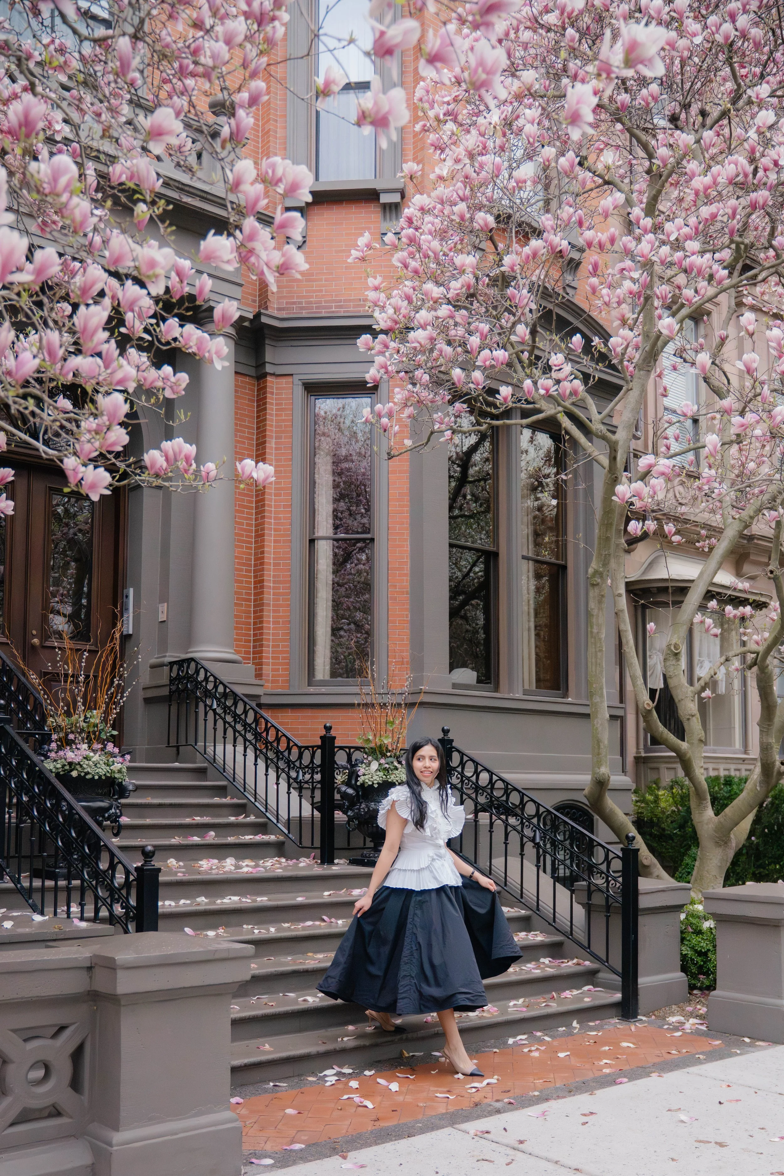 A woman in a white blouse and dark skirt walking down the steps of a city sidewalk, surrounded by blooming pink magnolia trees and a brownstone building with large windows.