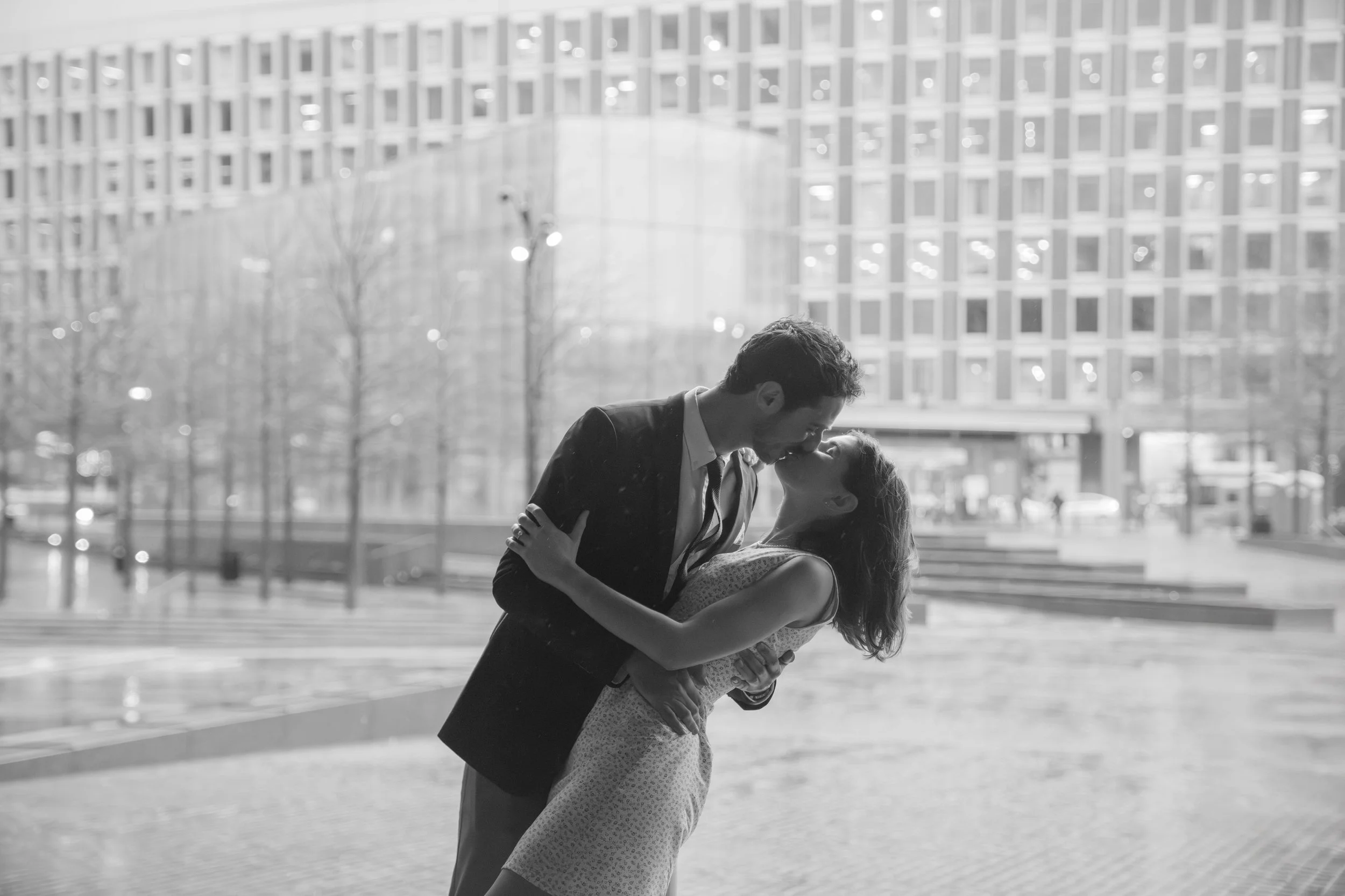 A couple sharing a kiss outdoors in front of a modern glass building, with the man in a suit and the woman in a dress.