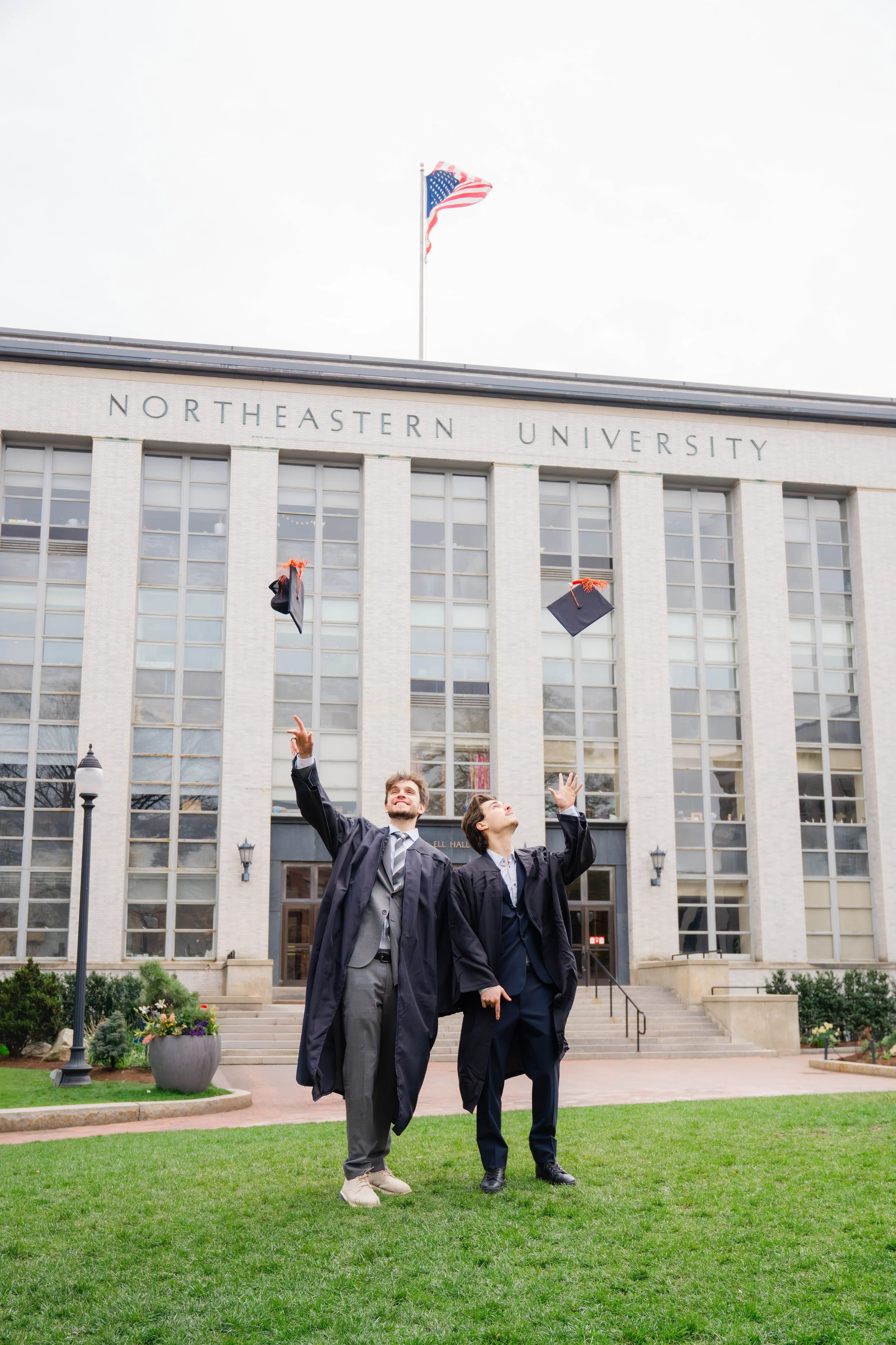 Two graduates in caps and gowns celebrating outside Northeastern University, tossing their caps into the air, with the university building and American flag in the background.