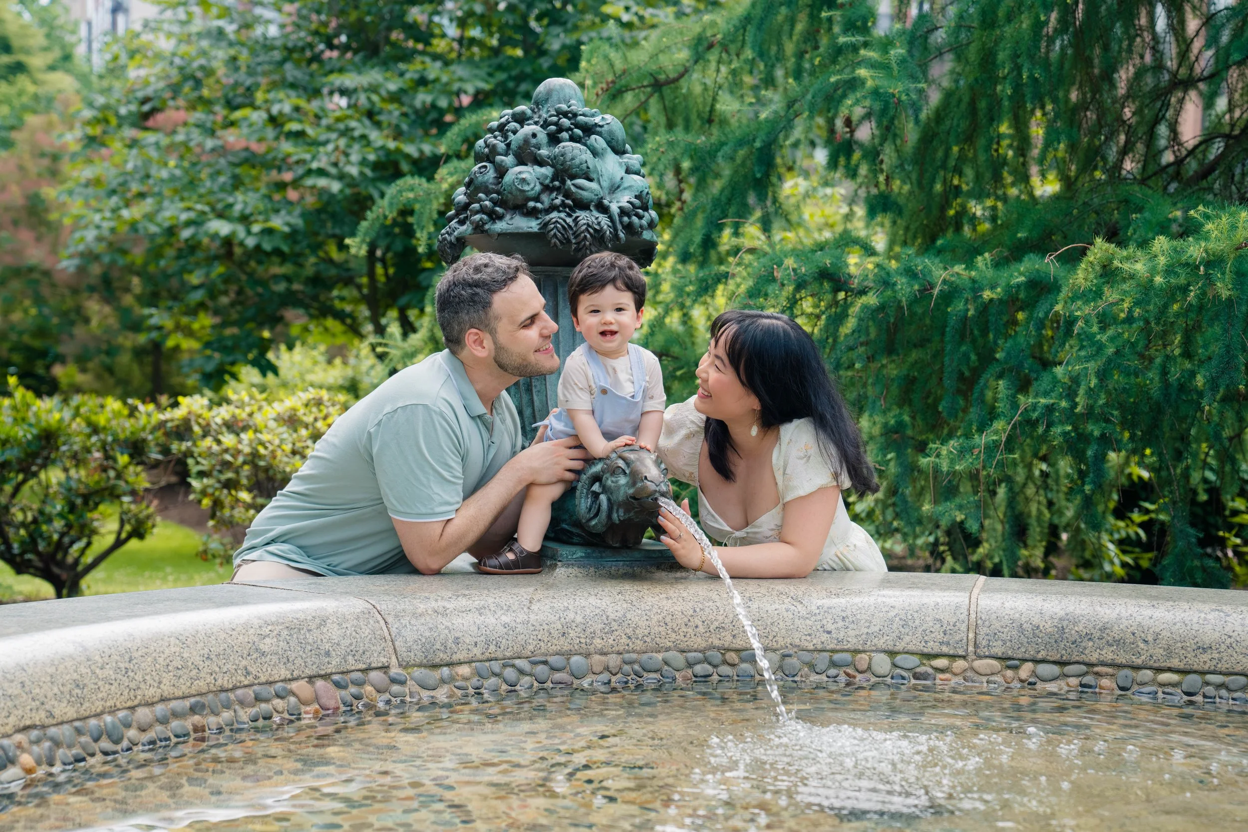 A family of three, including a man, woman, and young child, sitting by a decorative fountain in a lush garden with greenery.