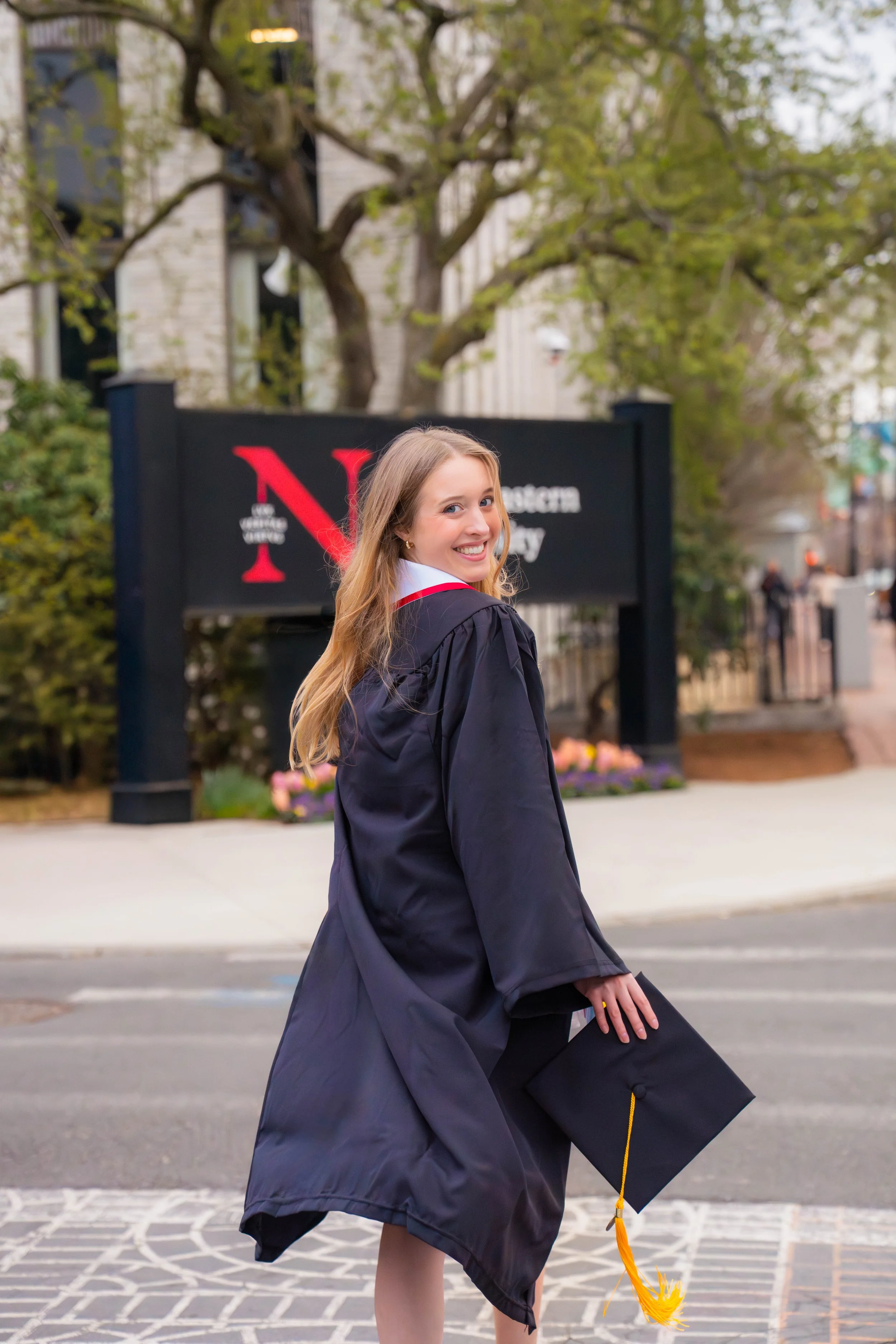 A young woman in a graduation gown and cap holding the cap in her hand, smiling and turning back while walking on a city street.