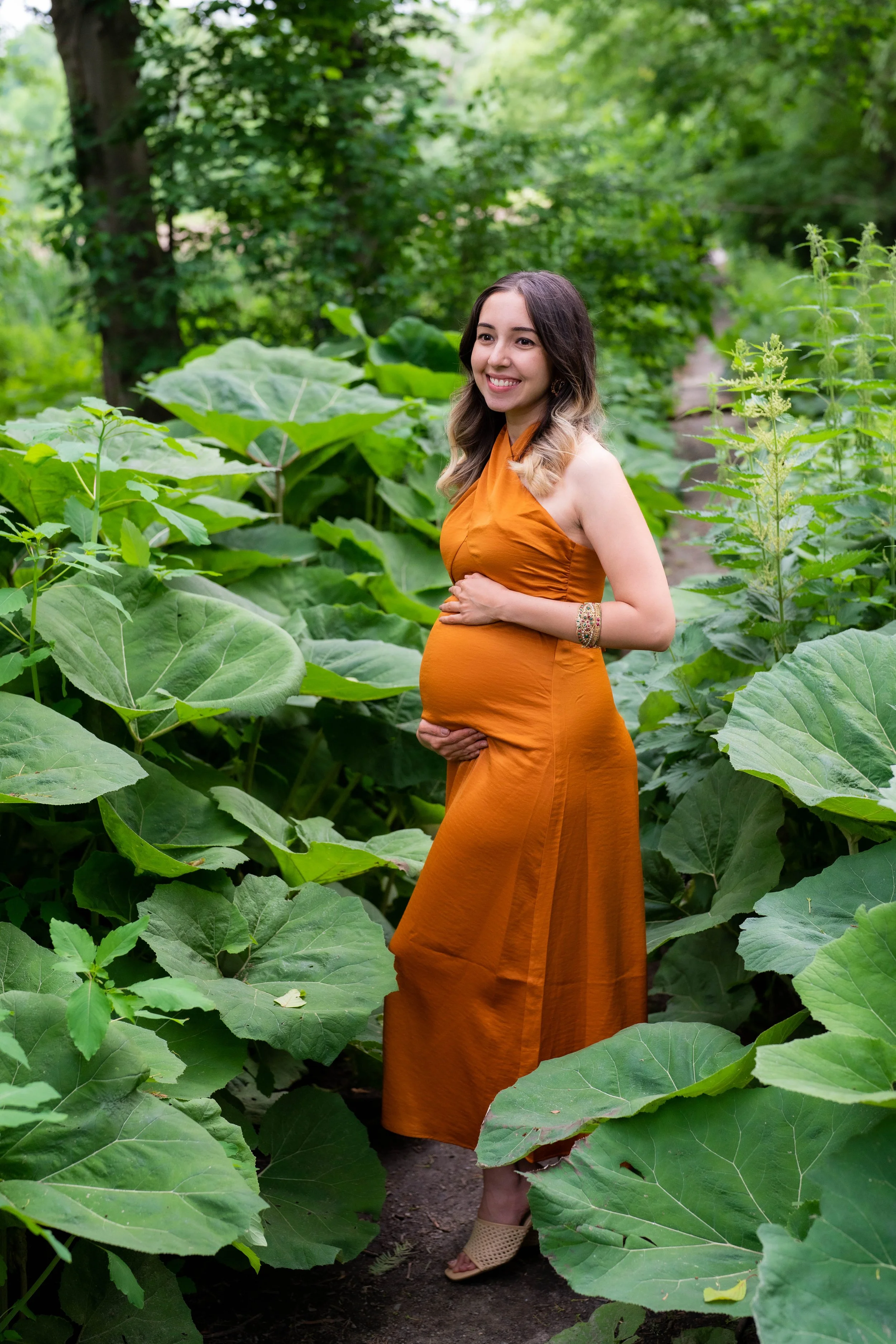 A pregnant woman smiling and standing among large green leaves in a lush, green outdoor setting.
