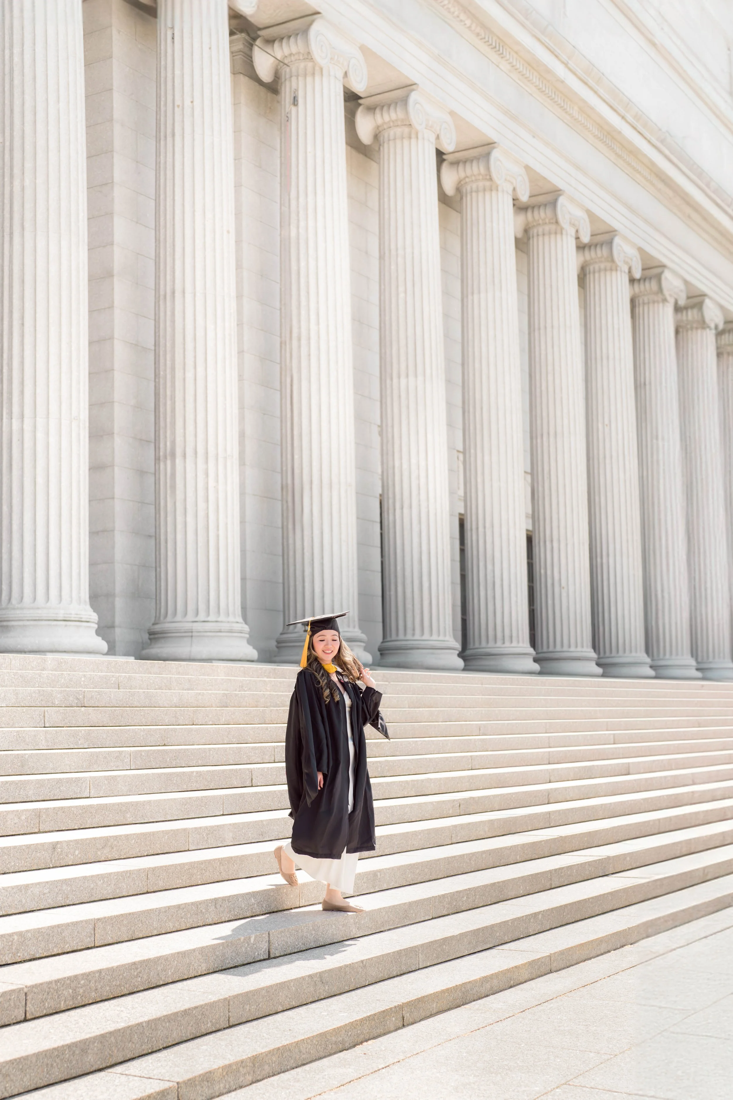 A young woman in a black graduation gown and mortarboard walking down the steps of a classical building with tall, white, fluted columns.