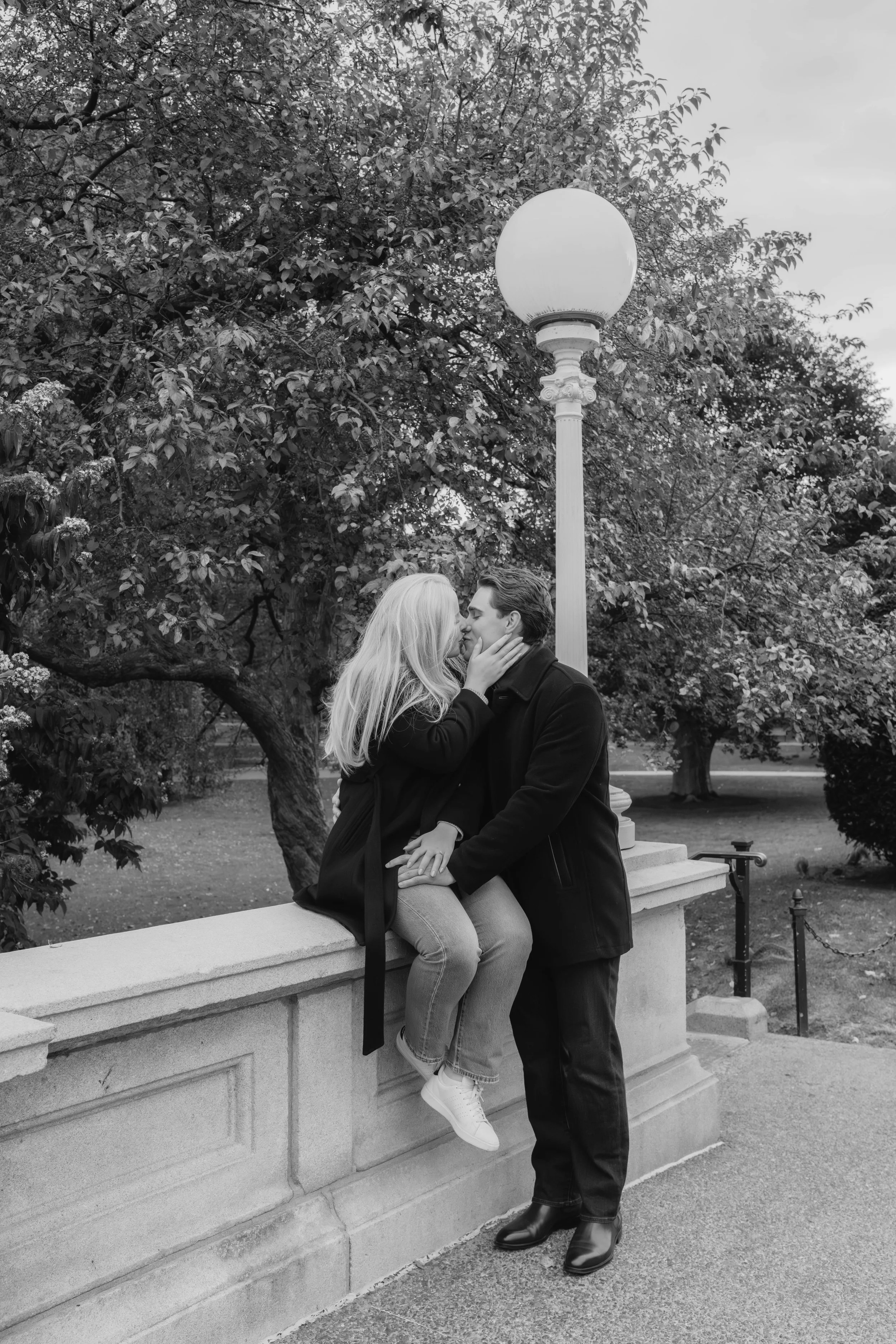A couple is kissing outdoors on a stone ledge, with the woman sitting on the ledge and the man standing in front of her. They are surrounded by trees and a streetlamp.