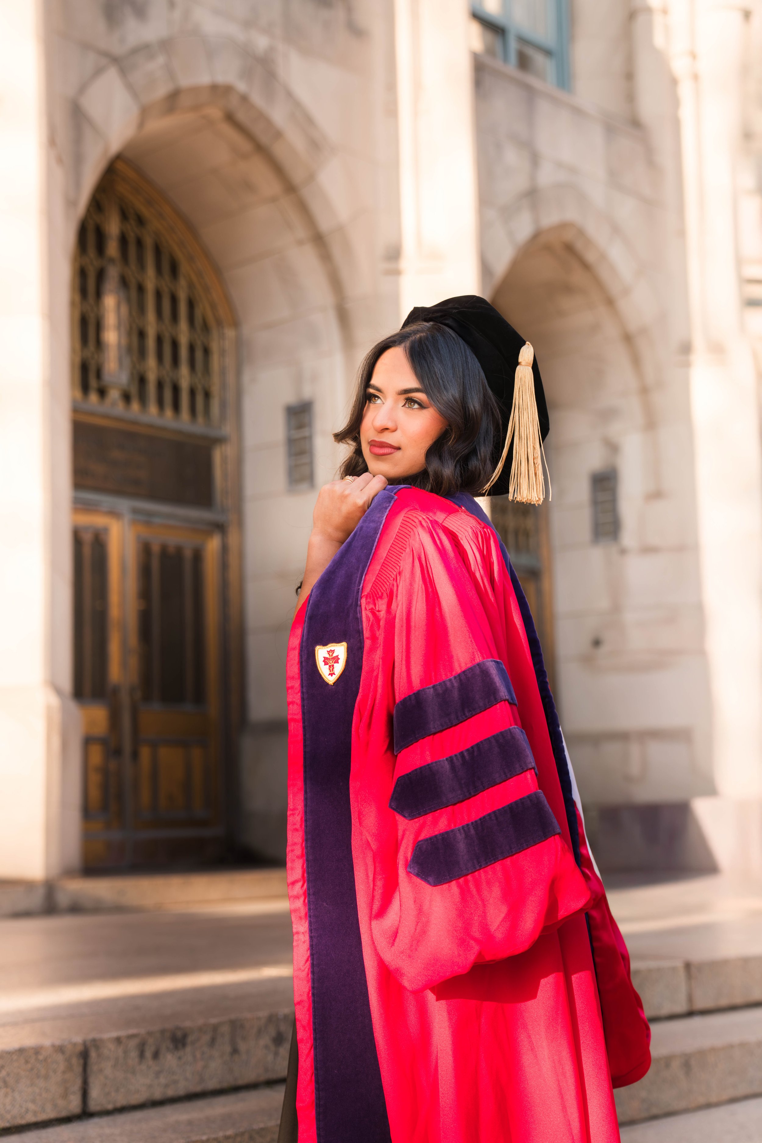 A young woman with dark hair in loose waves wearing a graduation cap with a gold tassel and a red academic gown. She stands outside in front of a historic building with stone arches and wooden doors, looking confidently into the distance.