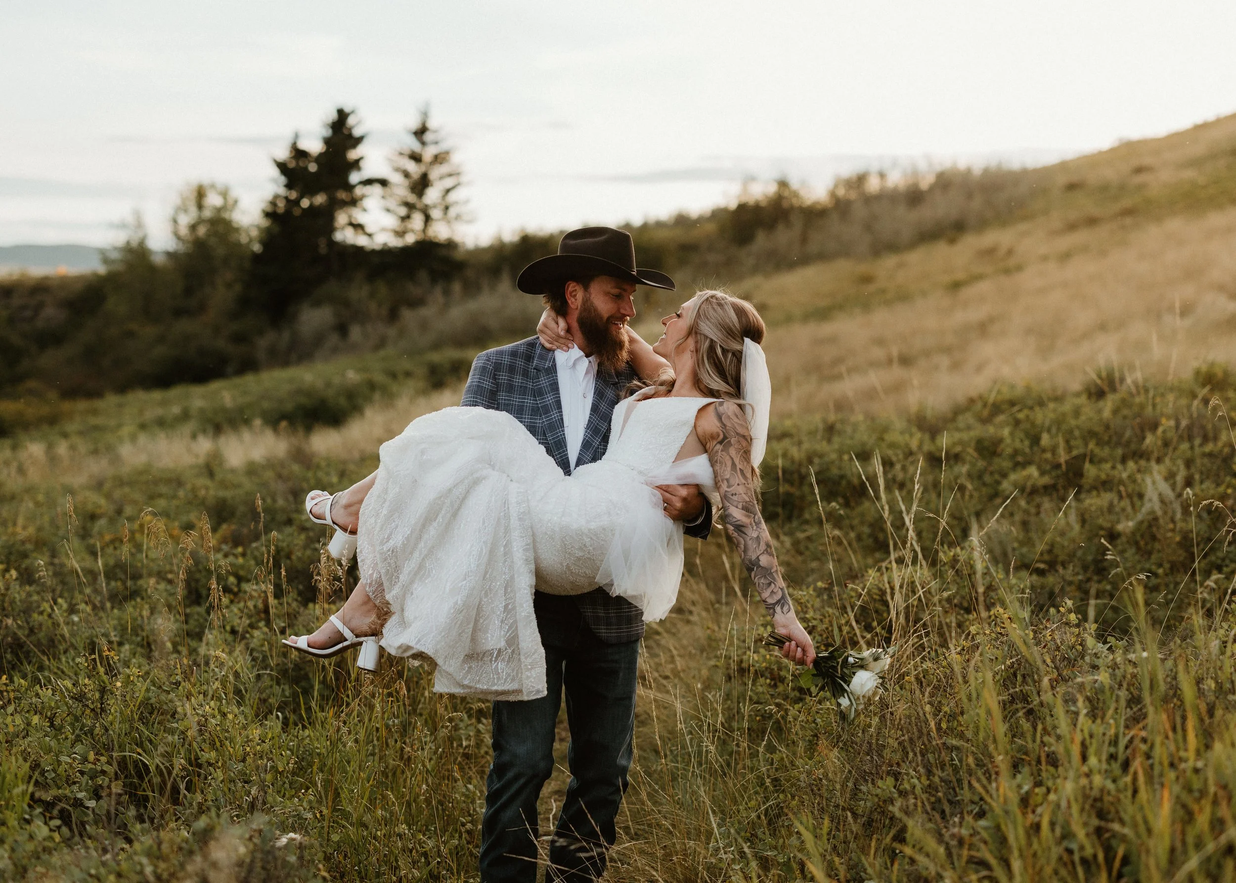 groom carries his bride through long grass in Cochrane, AB.