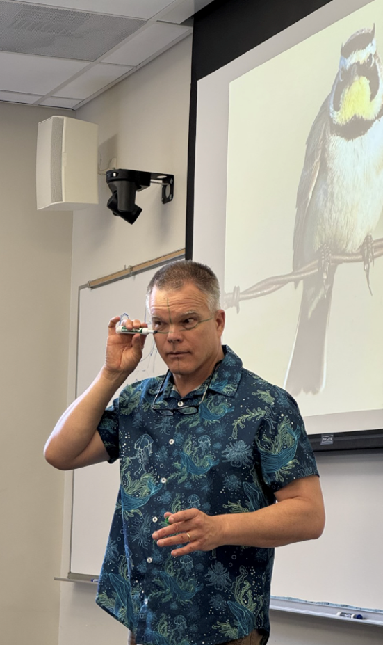 Jack Muir Laws in front of classroom and a slide of a bird, drawing planes on his face with a marker to illustrate sketching techniques.