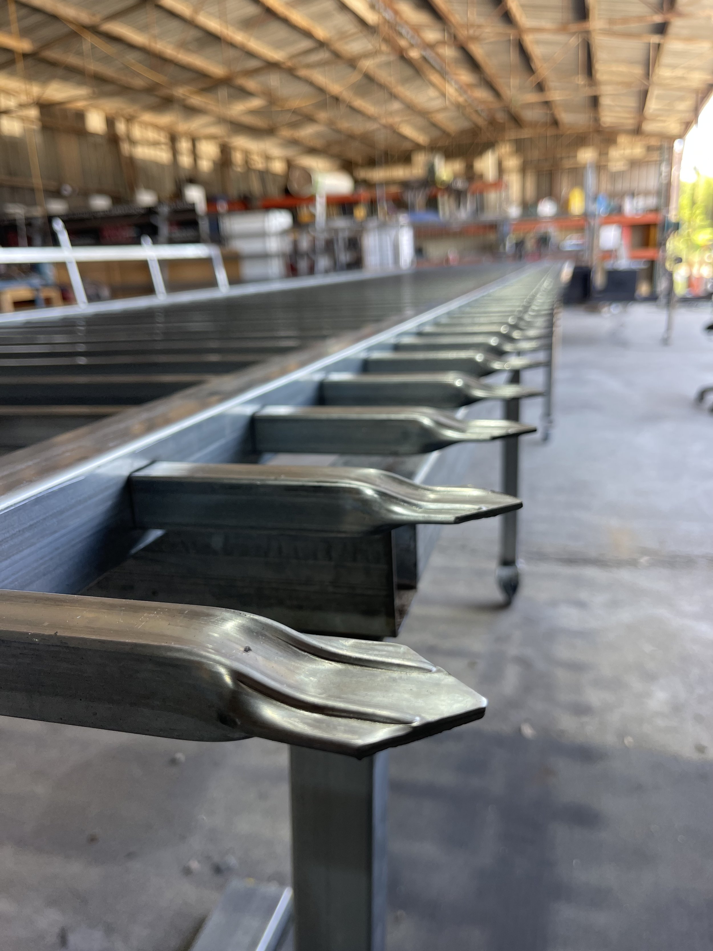 Close-up of a long, stainless steel bottle openers mounted on a metal table in a workshop or warehouse setting, with shelves and tools in the background.