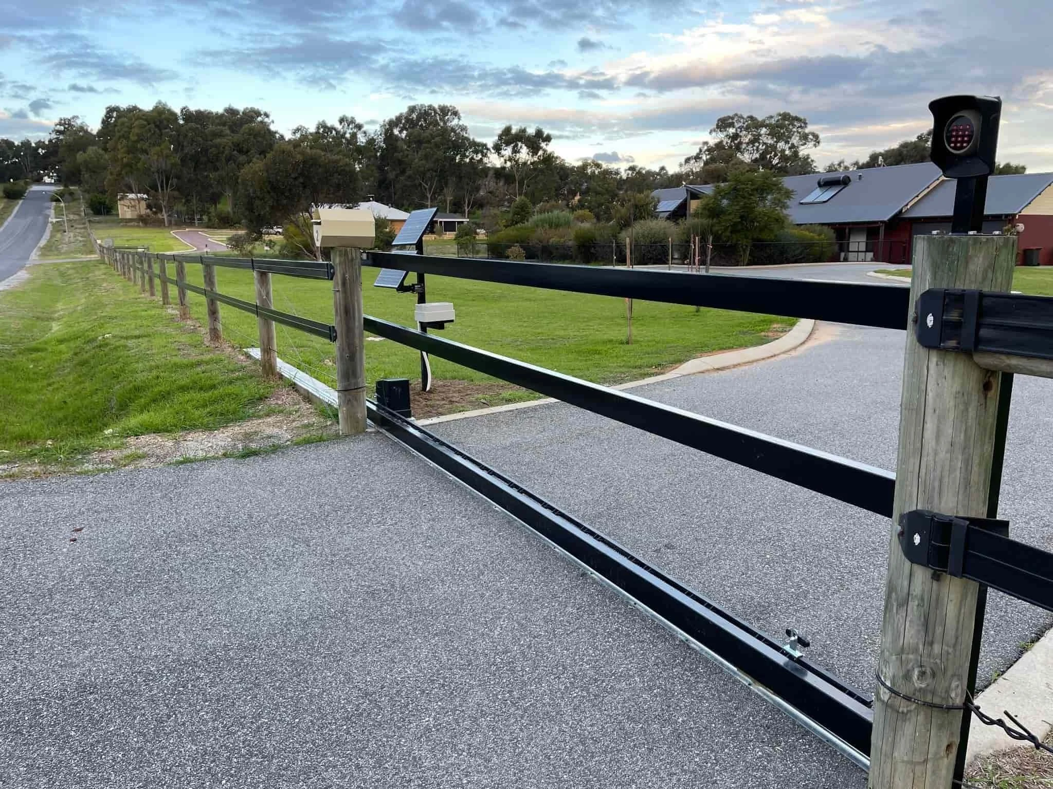 A driveway gate with solar panels, a mailbox, and a security camera, set in a suburban neighborhood with houses and trees in the background.