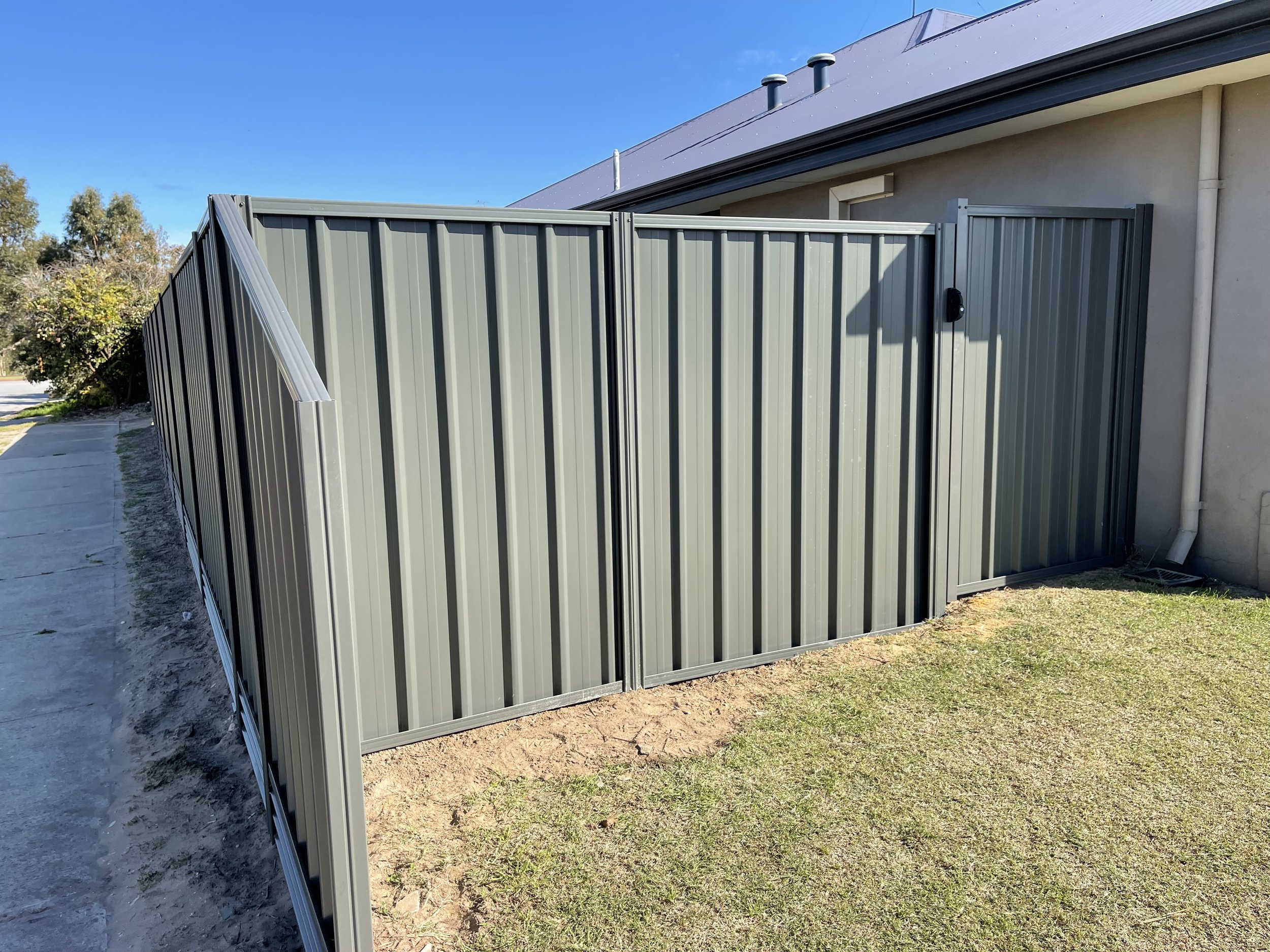 A gray metal privacy fence with a gate, installed around a house with a sloped roof and vent pipes, on a grassy yard under a clear blue sky.