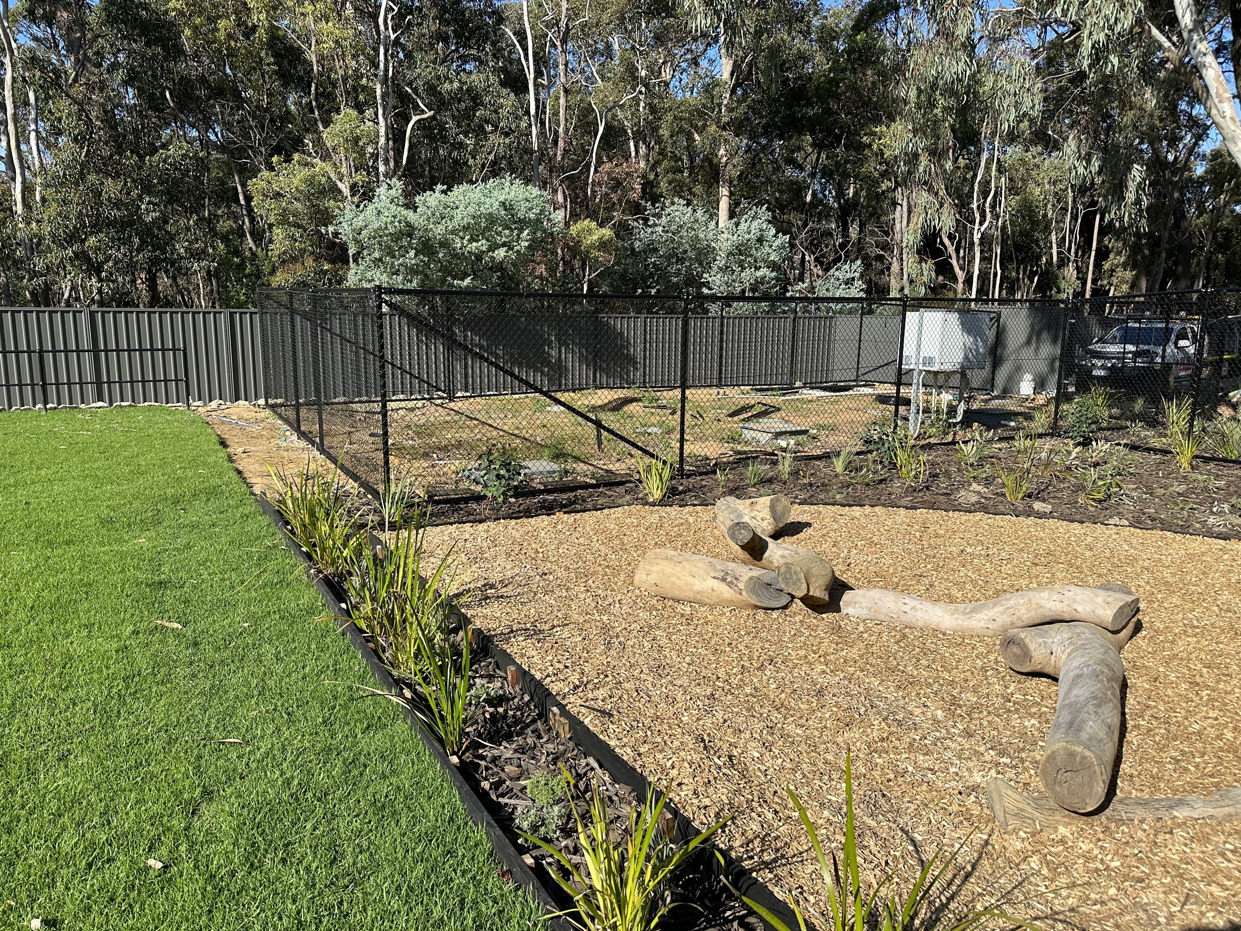 A backyard garden with green grass, a gravel area with wooden logs arranged as a seating or play area, young plants, and a black chain-link fence separating the garden from a wooded area with tall trees, a parked car, and utility boxes.