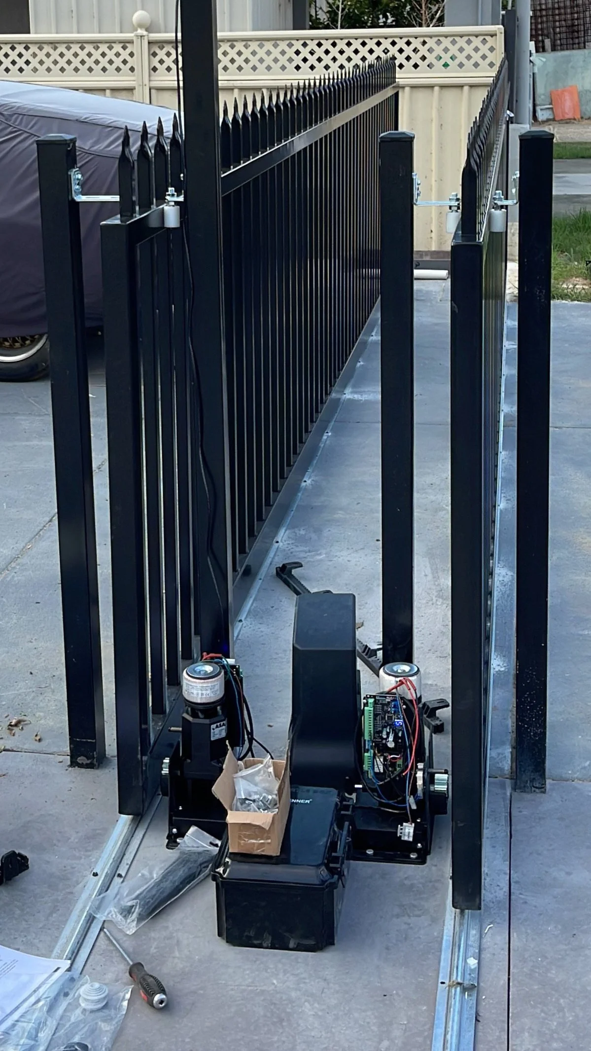 Black metal fence being assembled with an electric gate opener system and tools on a concrete driveway.