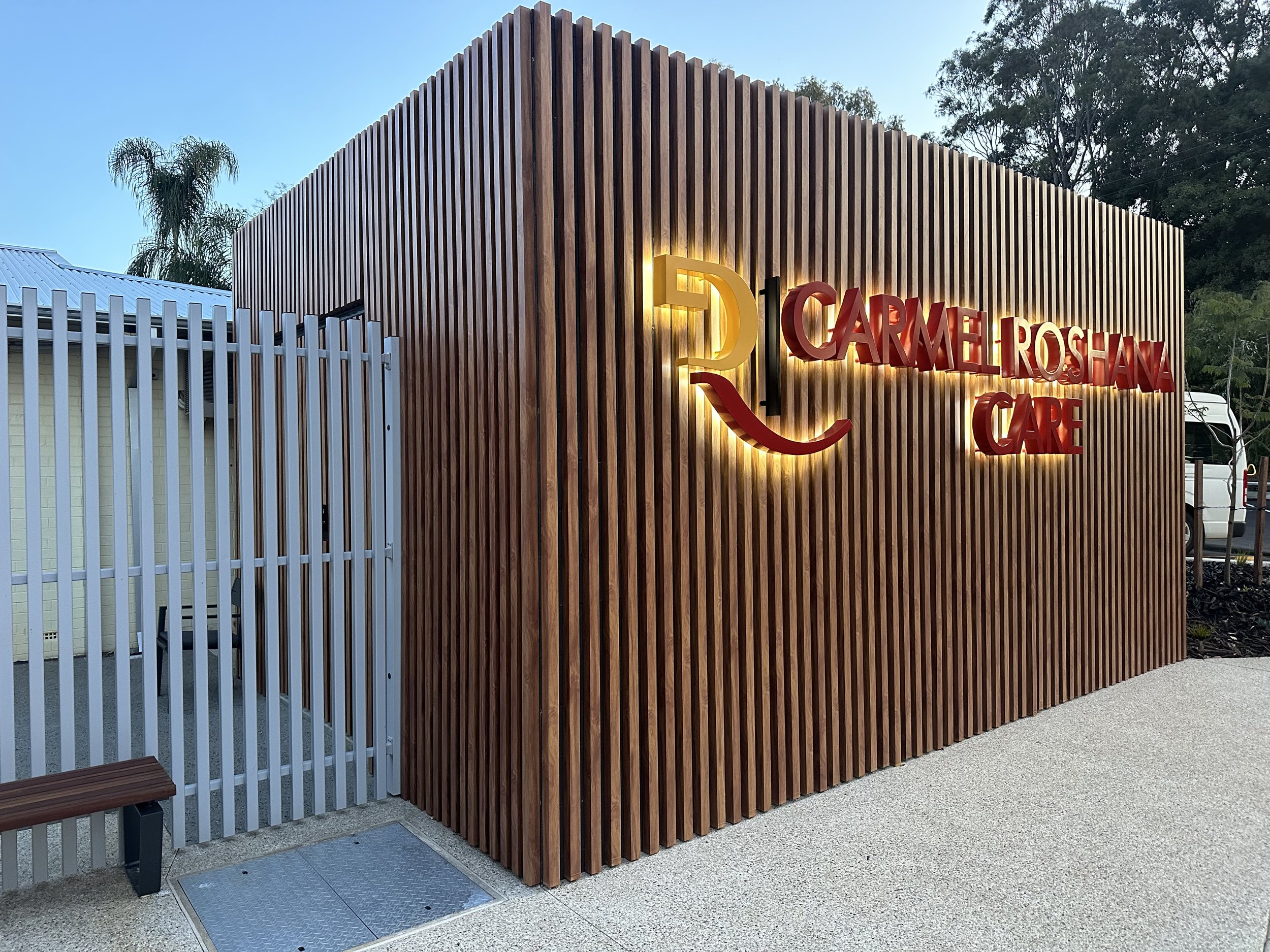 Exterior view of a modern wooden building with illuminated sign reading 'CARMEL ROSHANA CARE' and logo featuring a stylized face.