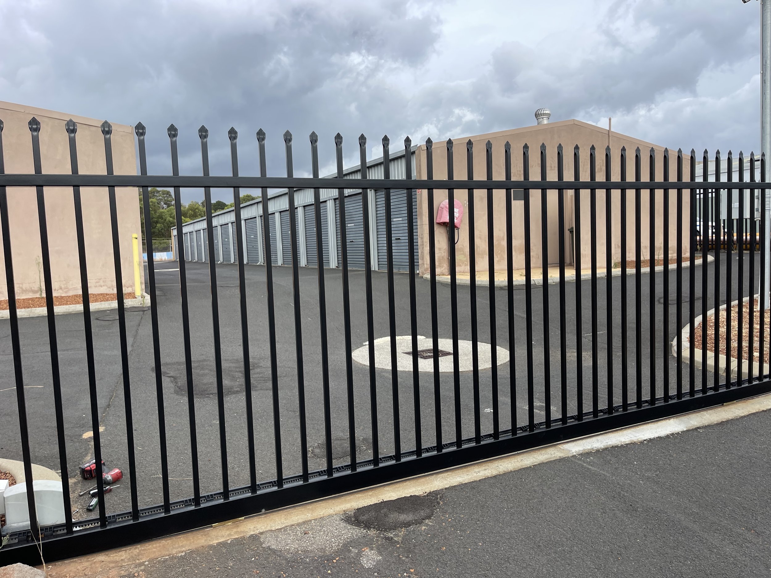 A black metal fence in front of a paved parking lot with closed storage unit doors and a beige building under a cloudy sky.