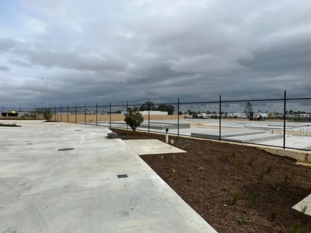 Empty parking lot with a chain-link fence, small trees, and cloudy sky.