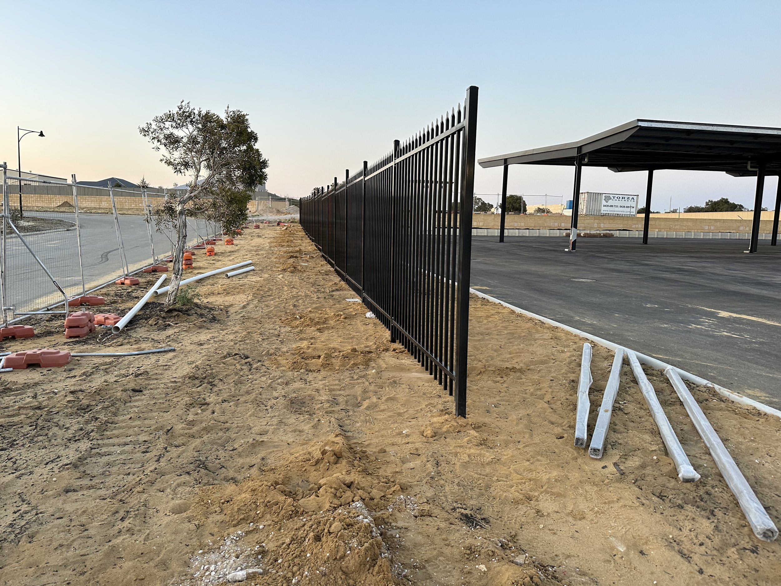 Construction site with a black metal fence, sandy ground, and a covered parking area in the background.