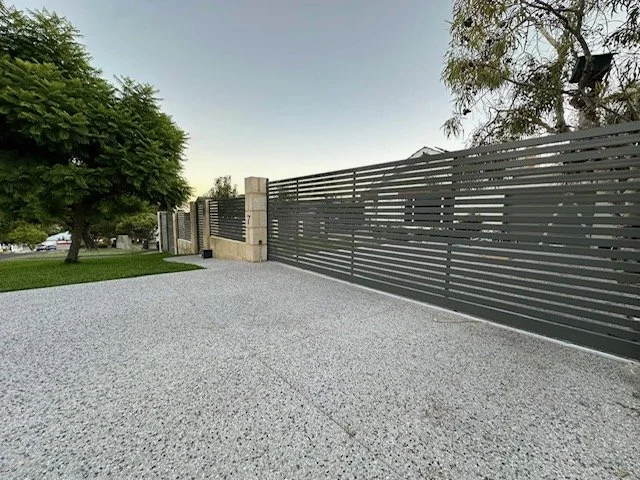 A modern black horizontal slatted metal fence runs along a light gray gravel driveway outside on a clear day, with green trees and a cloudy sky in the background.