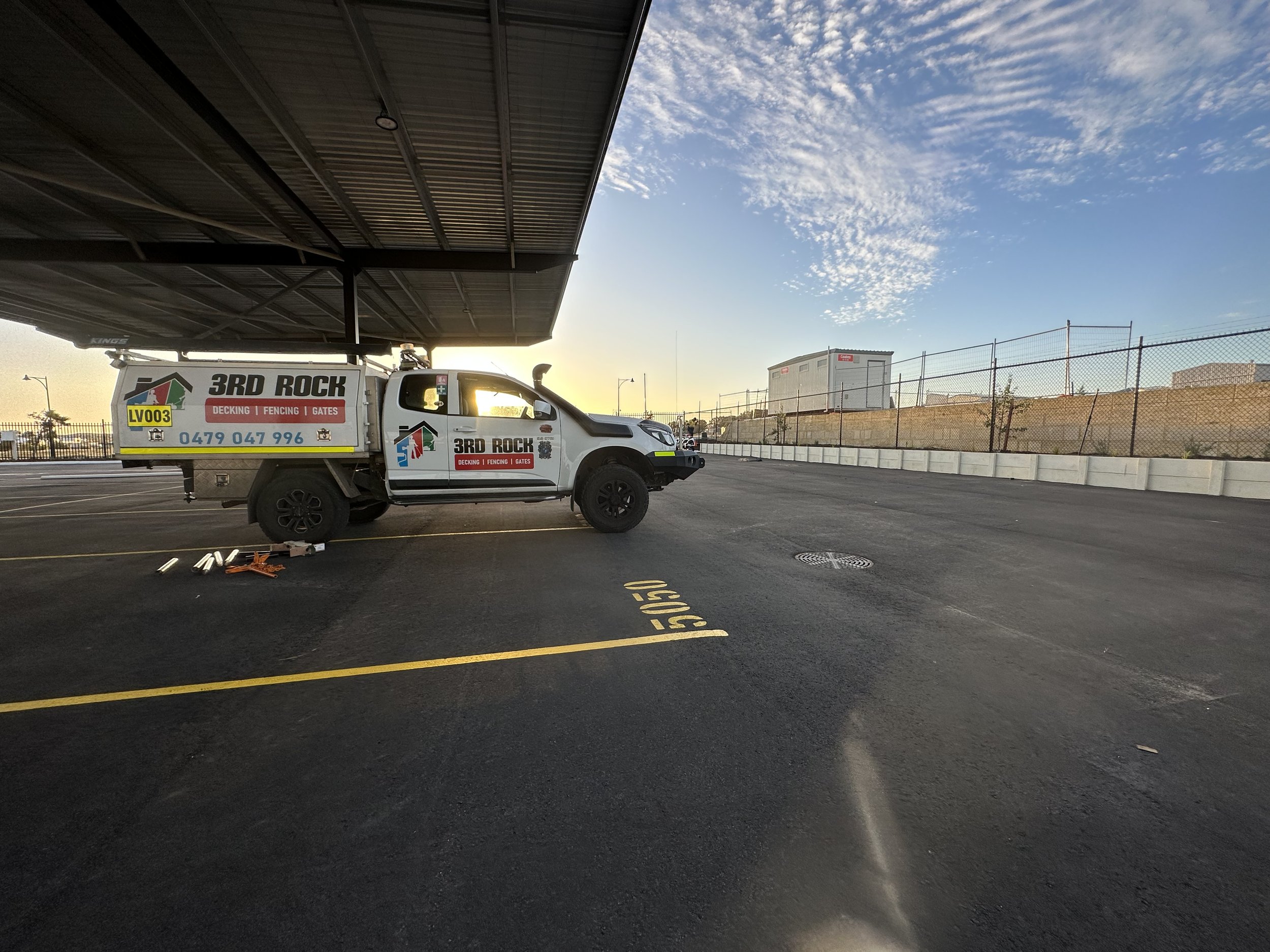 A white pickup truck with advertising for 3rd Rock, a fencing and gate company, is parked in a lot near a chain-link fence and an industrial building. The scene is captured during sunrise or sunset with clear skies.