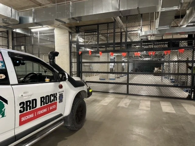 Part of a white pickup truck with 3RD ROCK branding parked in front of a security gate with traffic cones and fencing in an industrial or commercial parking area.