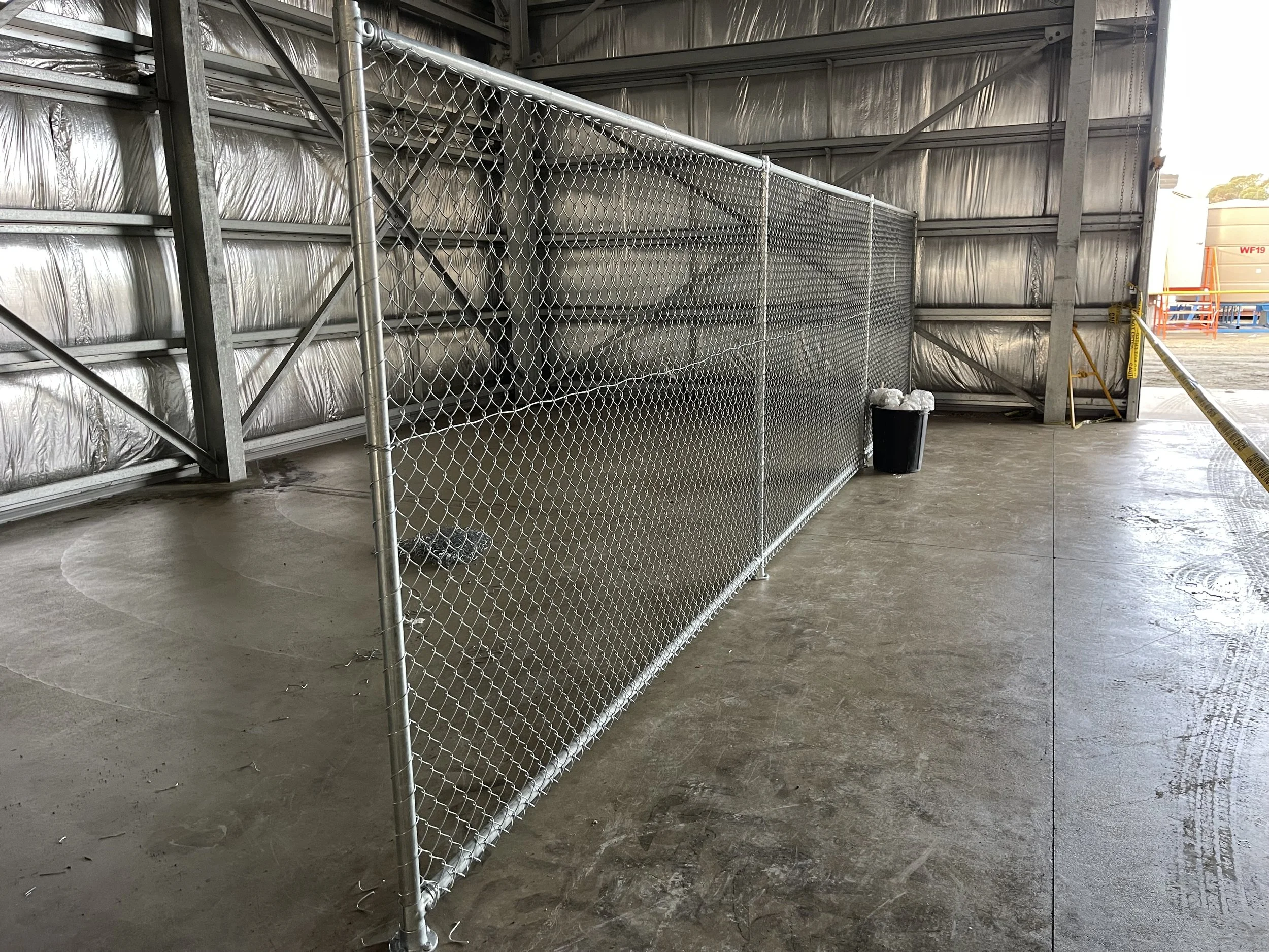 A chain-link fence in an industrial setting, with metal walls and concrete floor, near a trash can and yellow caution tape.