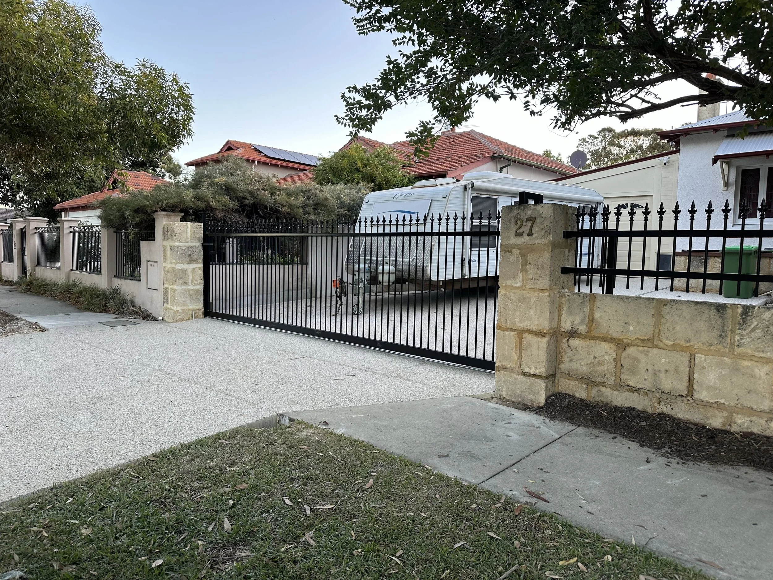 A residential front yard with a black metal gate and stone columns, a white trailer parked in drive, and houses with tiled roofs in the background.