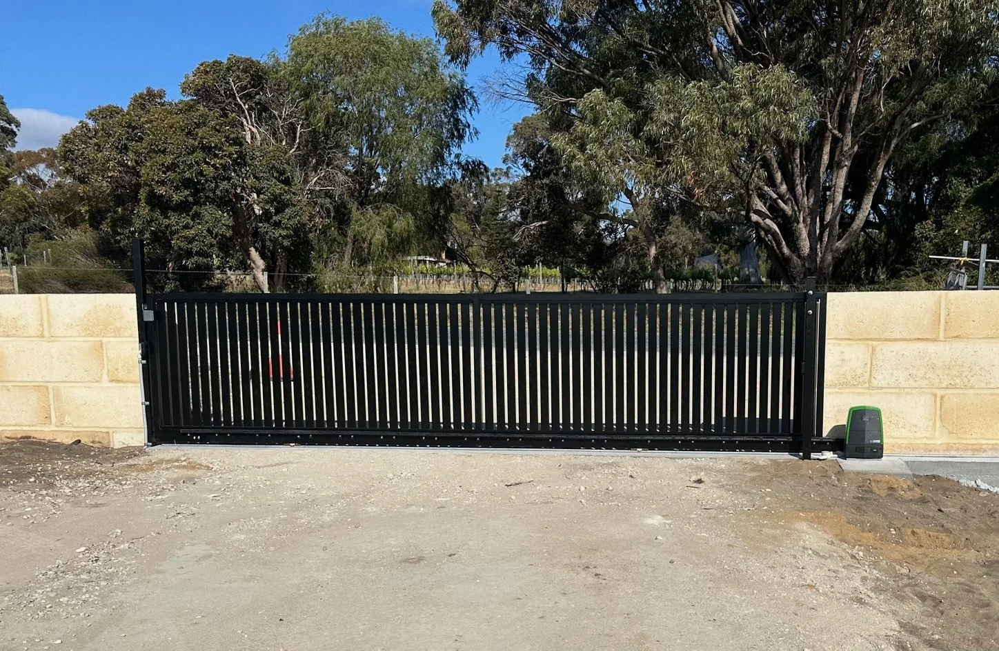 A black sliding gate installed in front of a beige brick wall, with trees and a clear blue sky in the background.