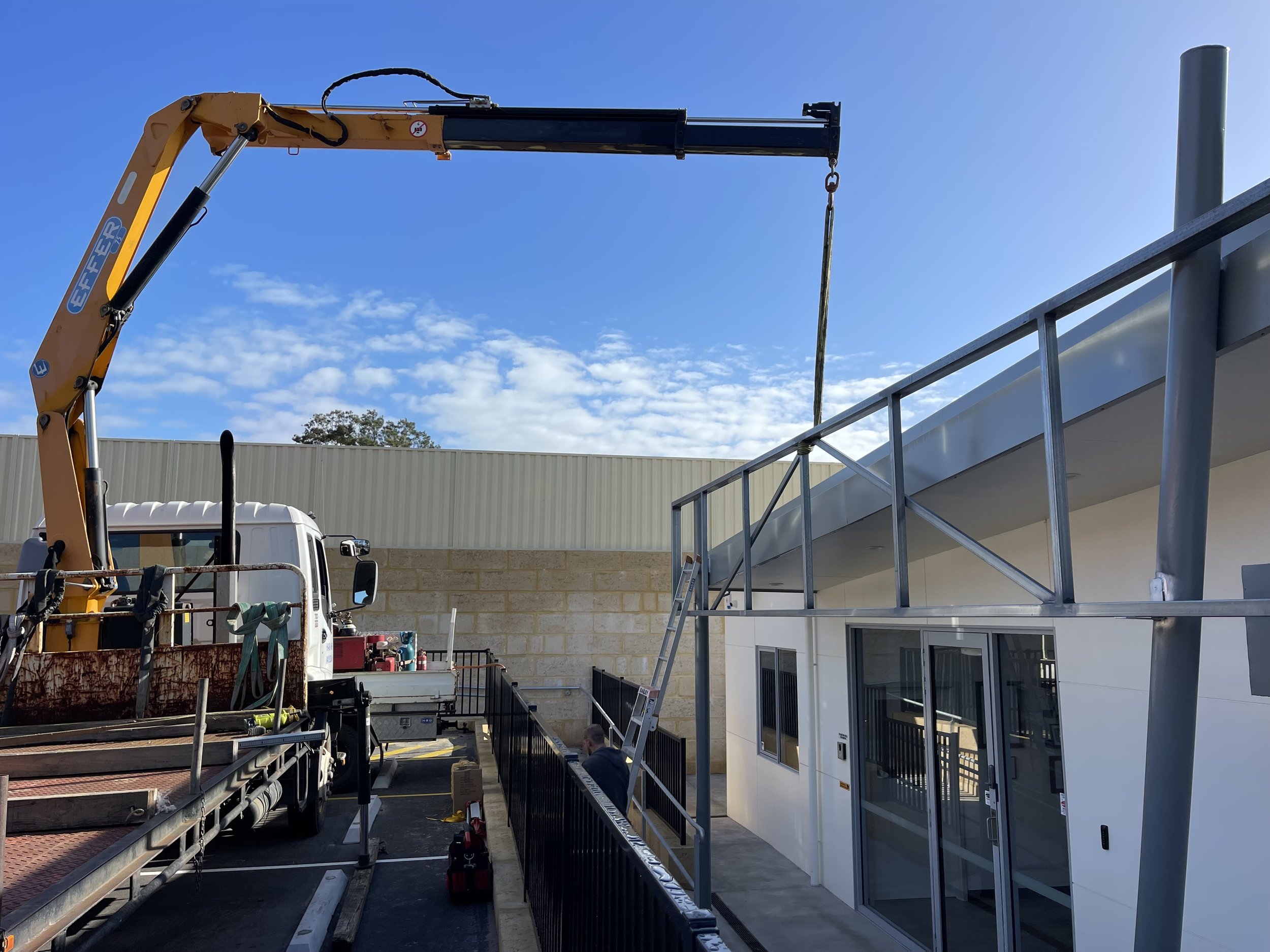 Construction site with a crane lifting a metal beam near a white building with glass doors and windows under a blue sky with some clouds.