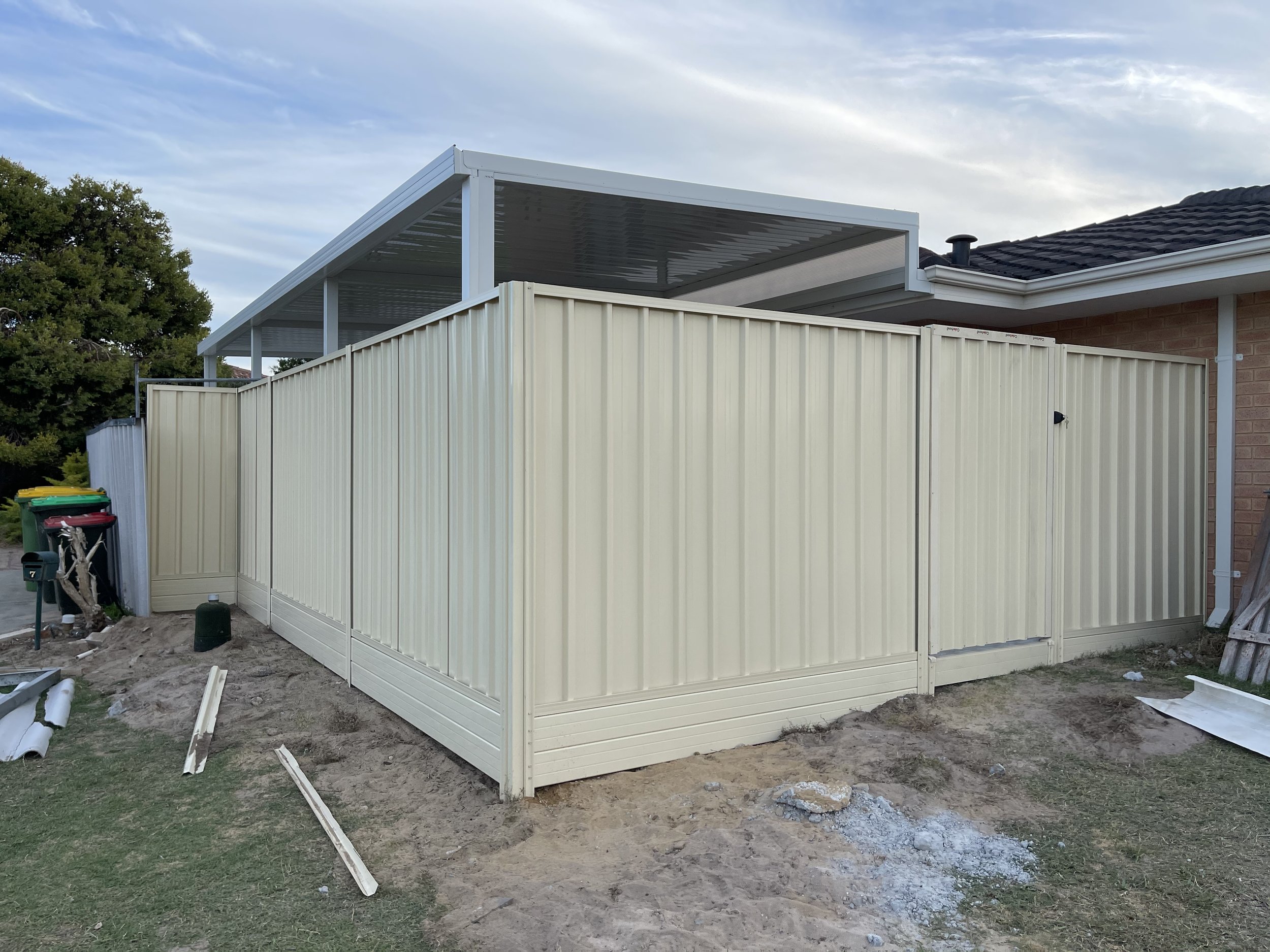 Exterior of a house with a beige metal fence and a partially constructed outdoor patio cover with a white metal roof.