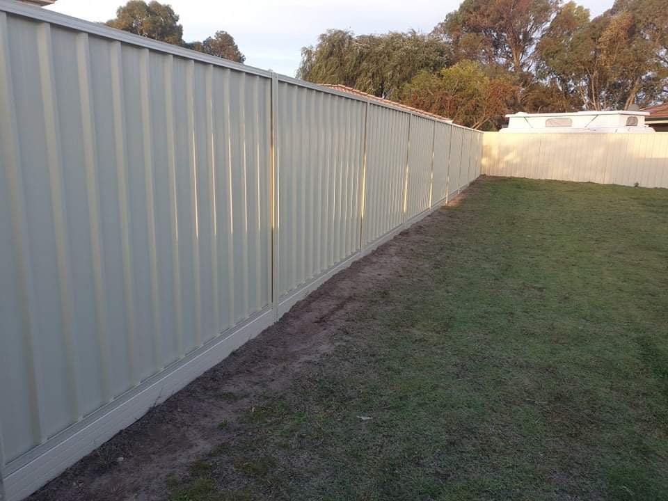A beige metal fence running along a grassy backyard with trees in the background.