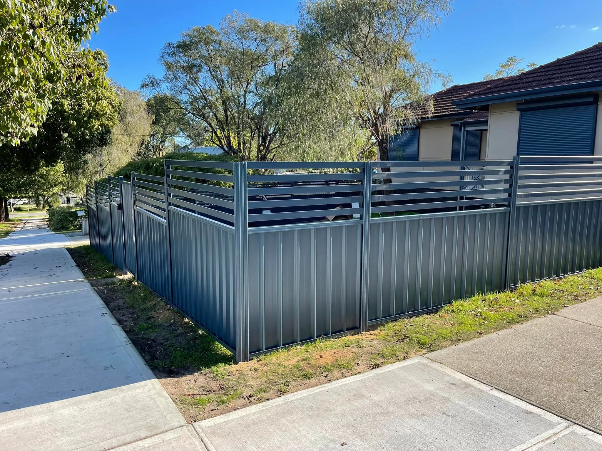 A metal fence with horizontal slats surrounds a corner of a house, with a sidewalk and grass in front and trees in the background.