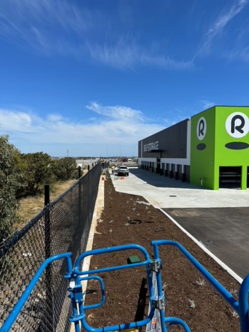 View of a shopping area with a large green and black retail store, parking lot with a few cars, and a playground structure in the foreground, under a partly cloudy sky.