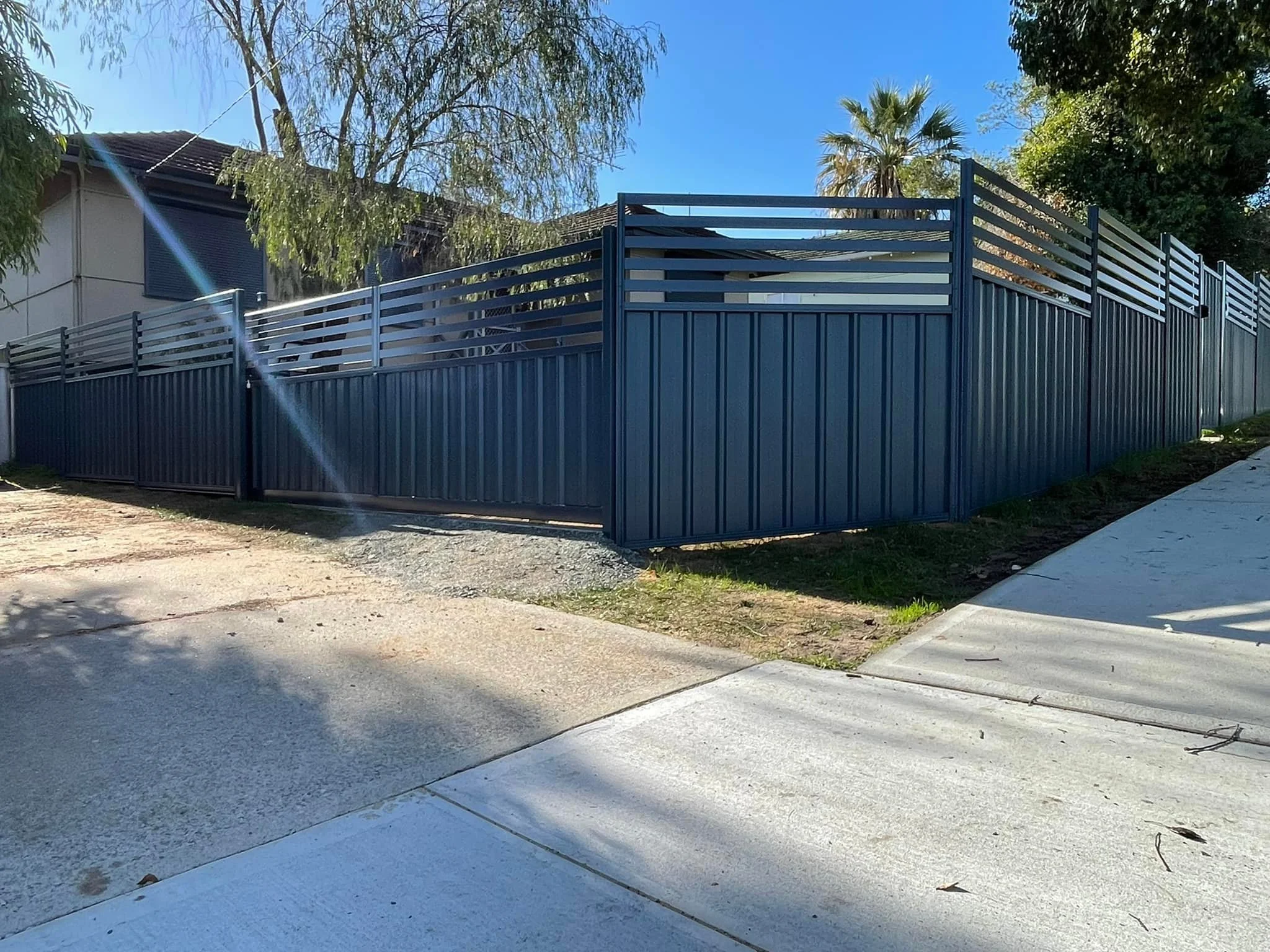 A tall dark blue metal fence with horizontal slats on the top and vertical slats on the bottom, enclosing a residential yard under a clear blue sky. There is a house partially visible behind the fence, several trees, and a sidewalk in the foreground.