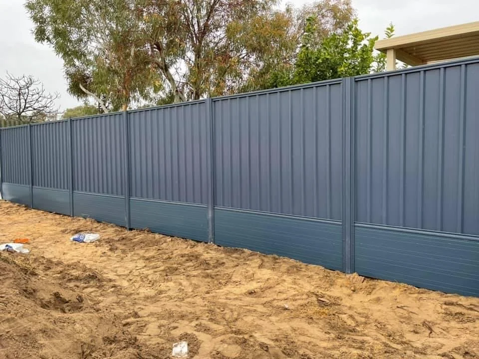 A metal fence painted in blue with vertical and horizontal panels, set on sandy ground with some trash debris, and trees in the background.