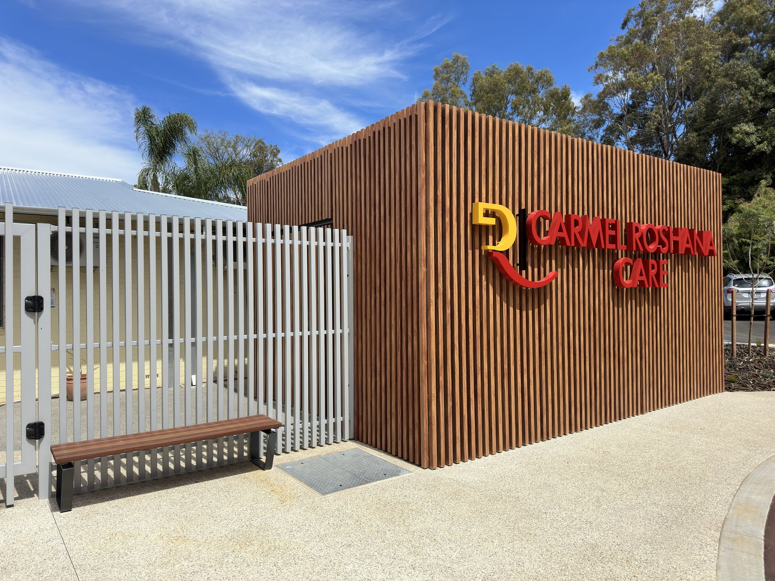 Exterior of Carmel Roshana Care facility with a wooden textured wall and colorful signage under a blue sky with clouds