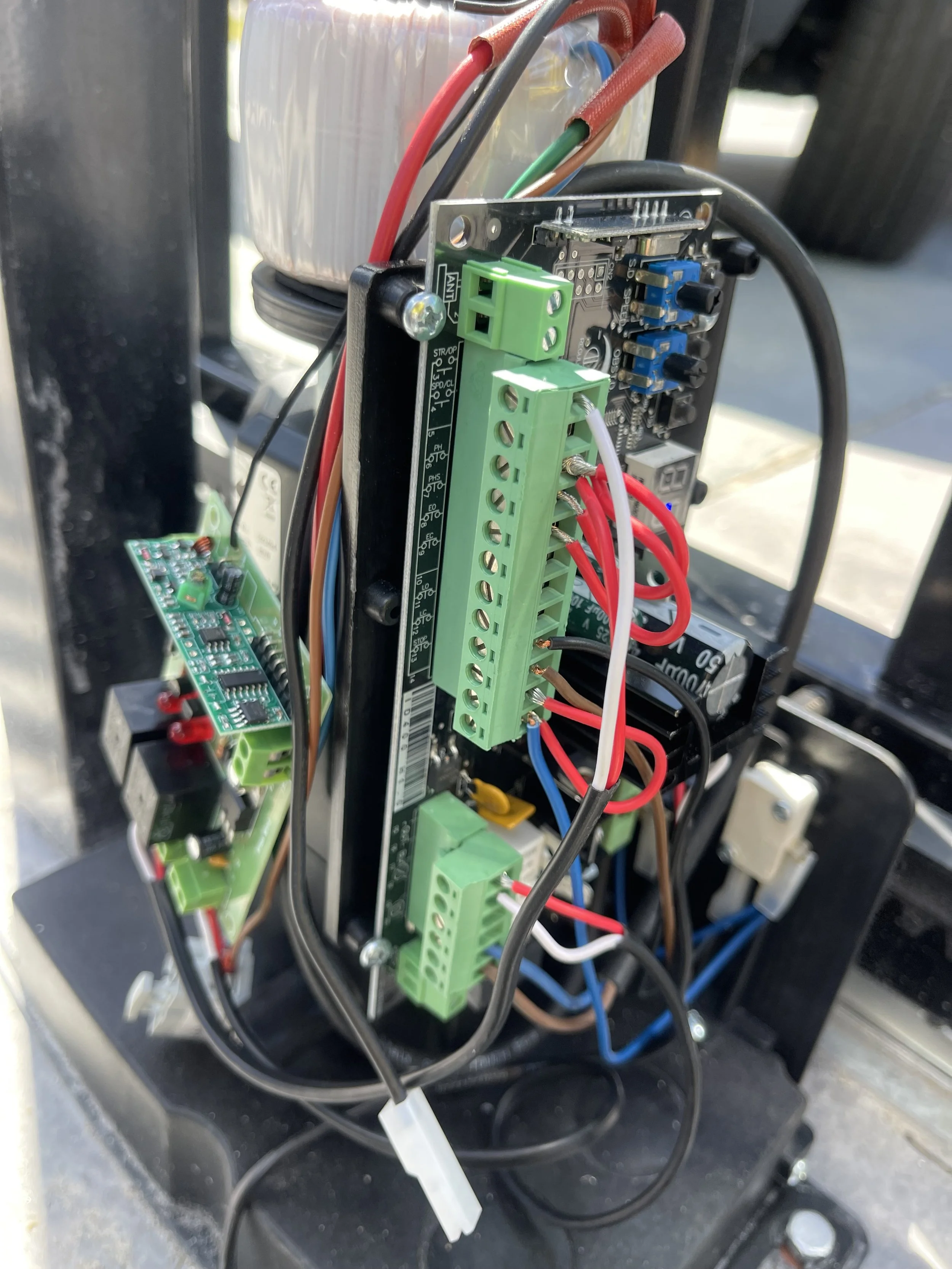 Close-up of an electronic control panel with green terminal blocks, circuit boards, and various wires connected, mounted on a black metal frame.
