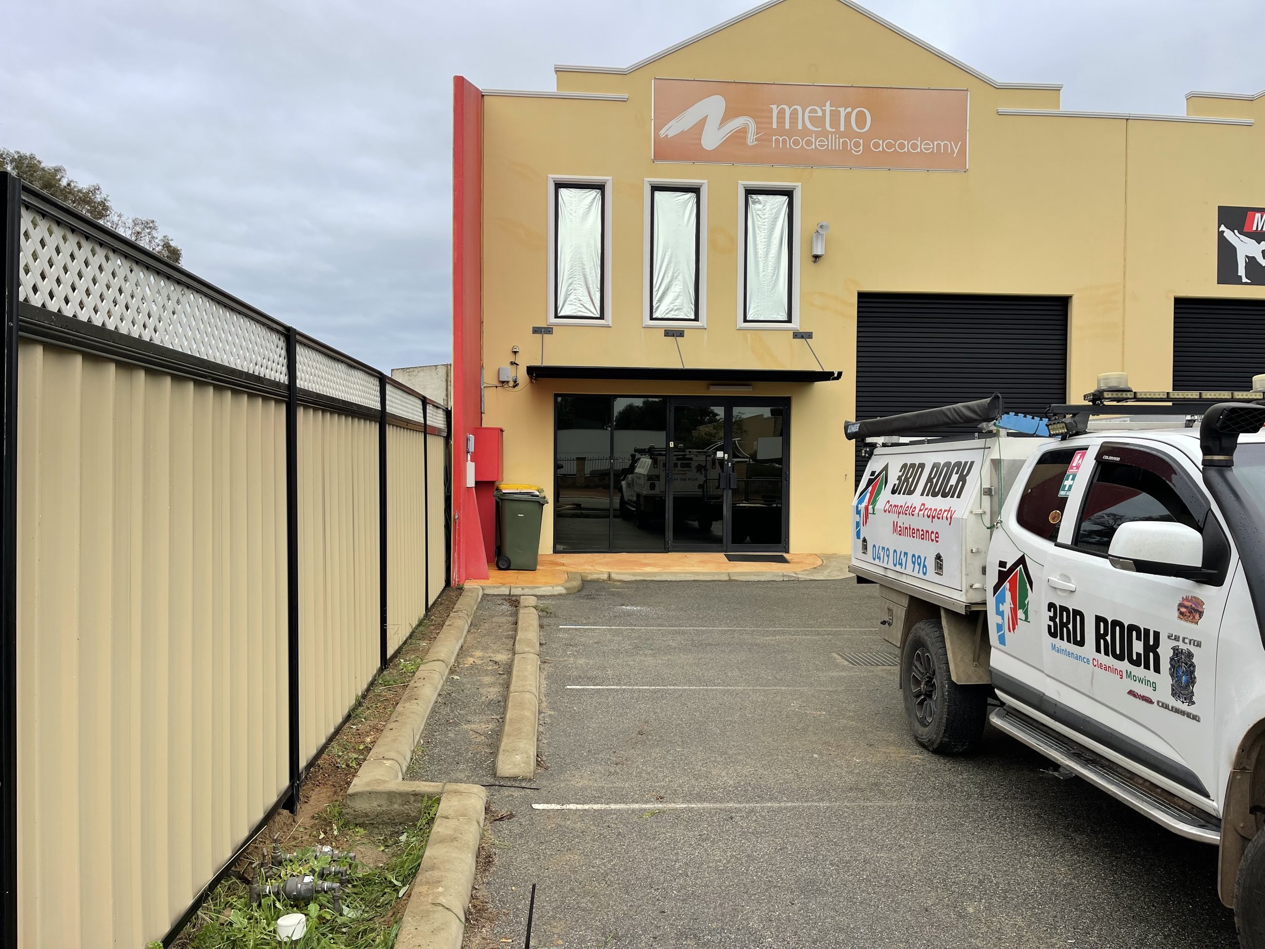 Front of a yellow building with a sign that reads 'metro modelling academy,' a glass entrance door, and three windows covered with aluminum foil, with a parked truck bearing a business logo and contact number in the parking lot.