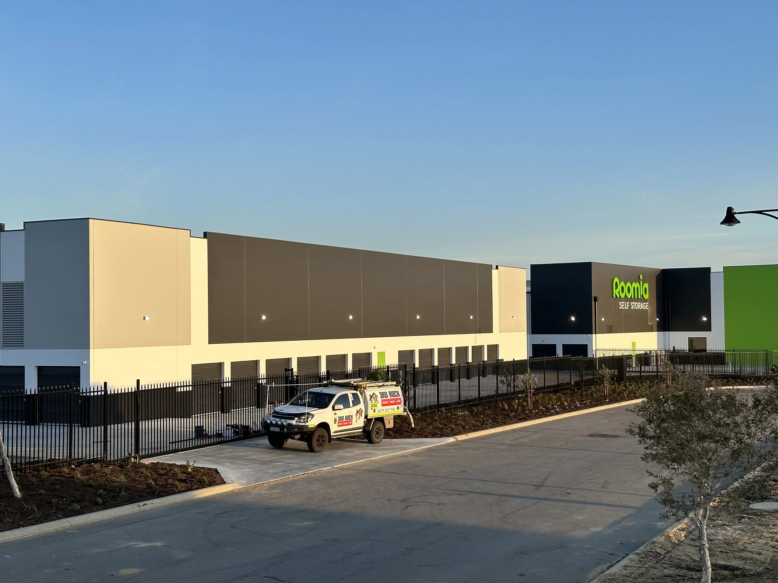 Exterior of a Roomia self-storage facility with a parked white vehicle in front, bordered by a black fence and landscaped areas, under a clear blue sky.
